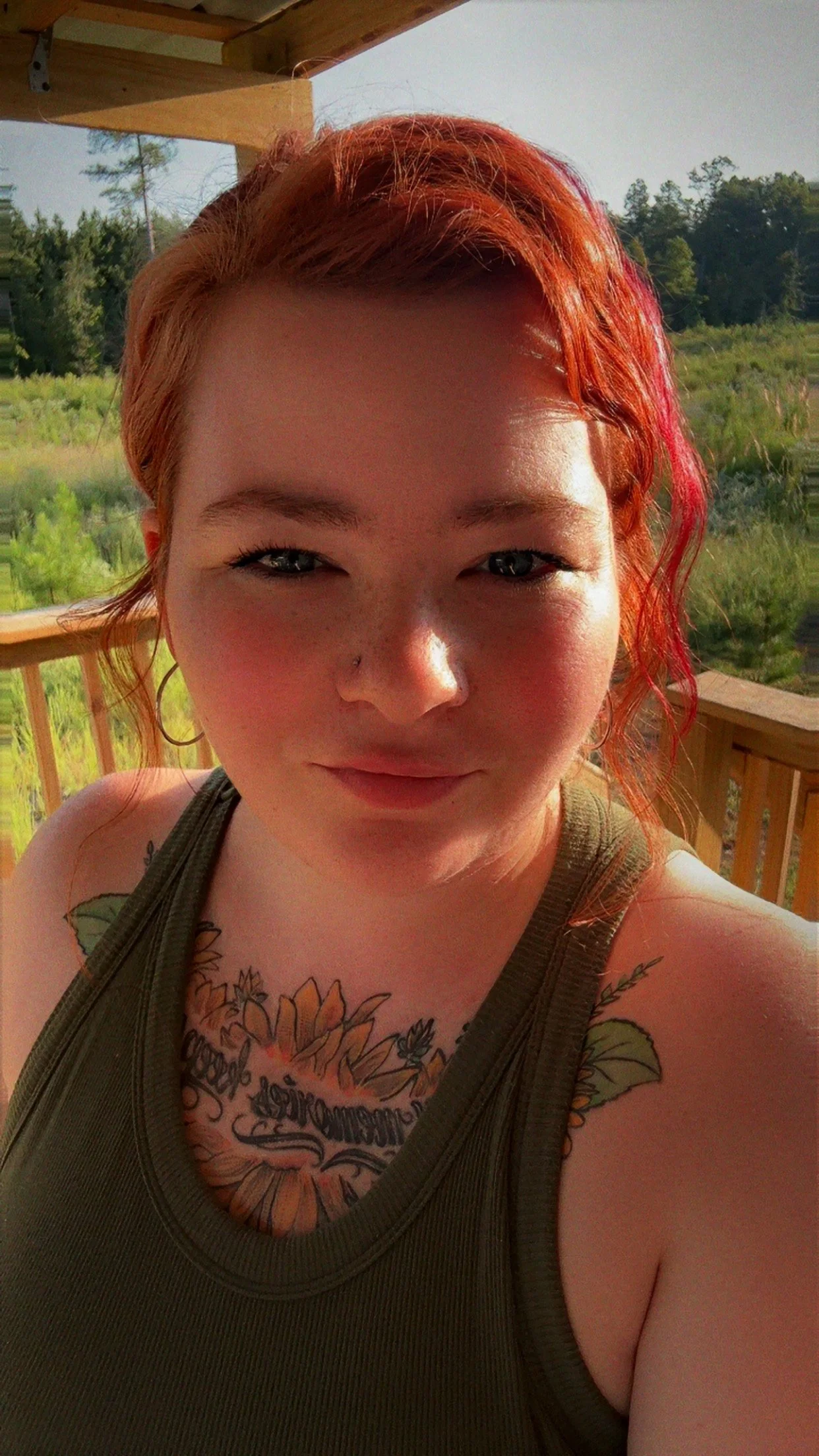 A woman with red hair and a nose piercing taking a selfie outdoors on a wooden deck, with a green landscape in the background.