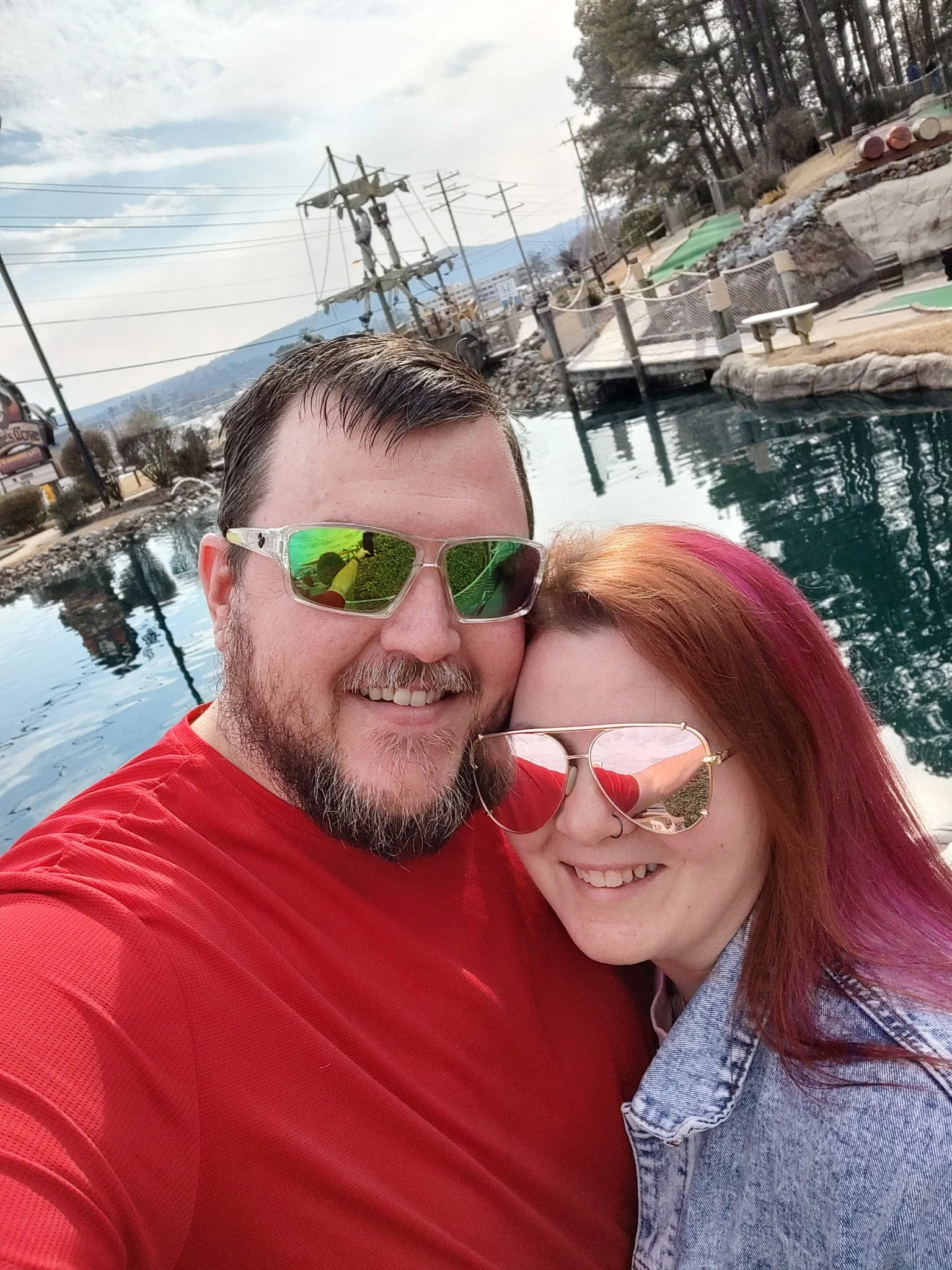 A smiling man and woman taking a selfie in front of a pool with a pirate ship and trees in the background.