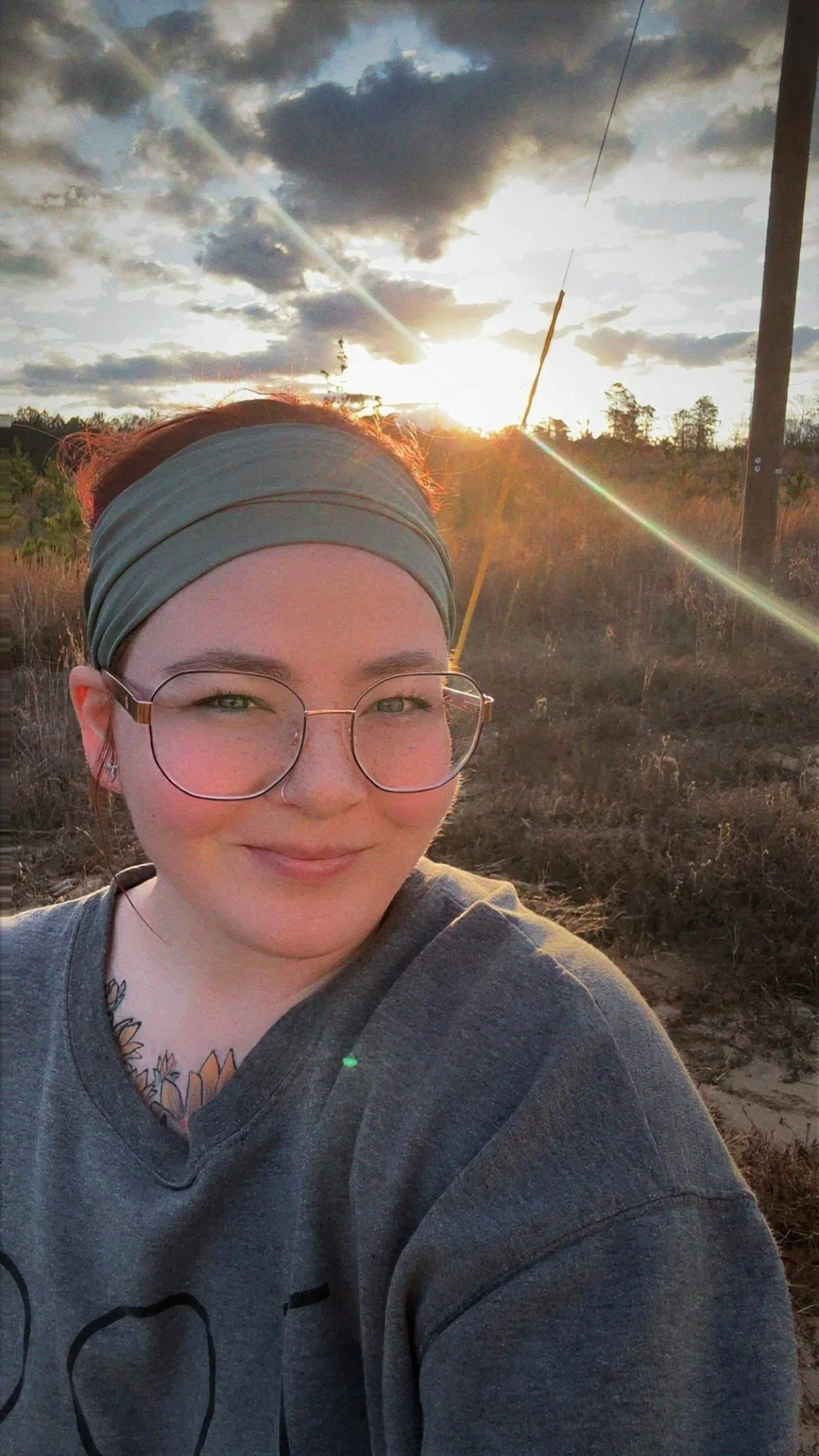 A young woman with glasses and a headband taking a selfie outdoors during sunset in a rural area, with power lines and dry vegetation in the background.