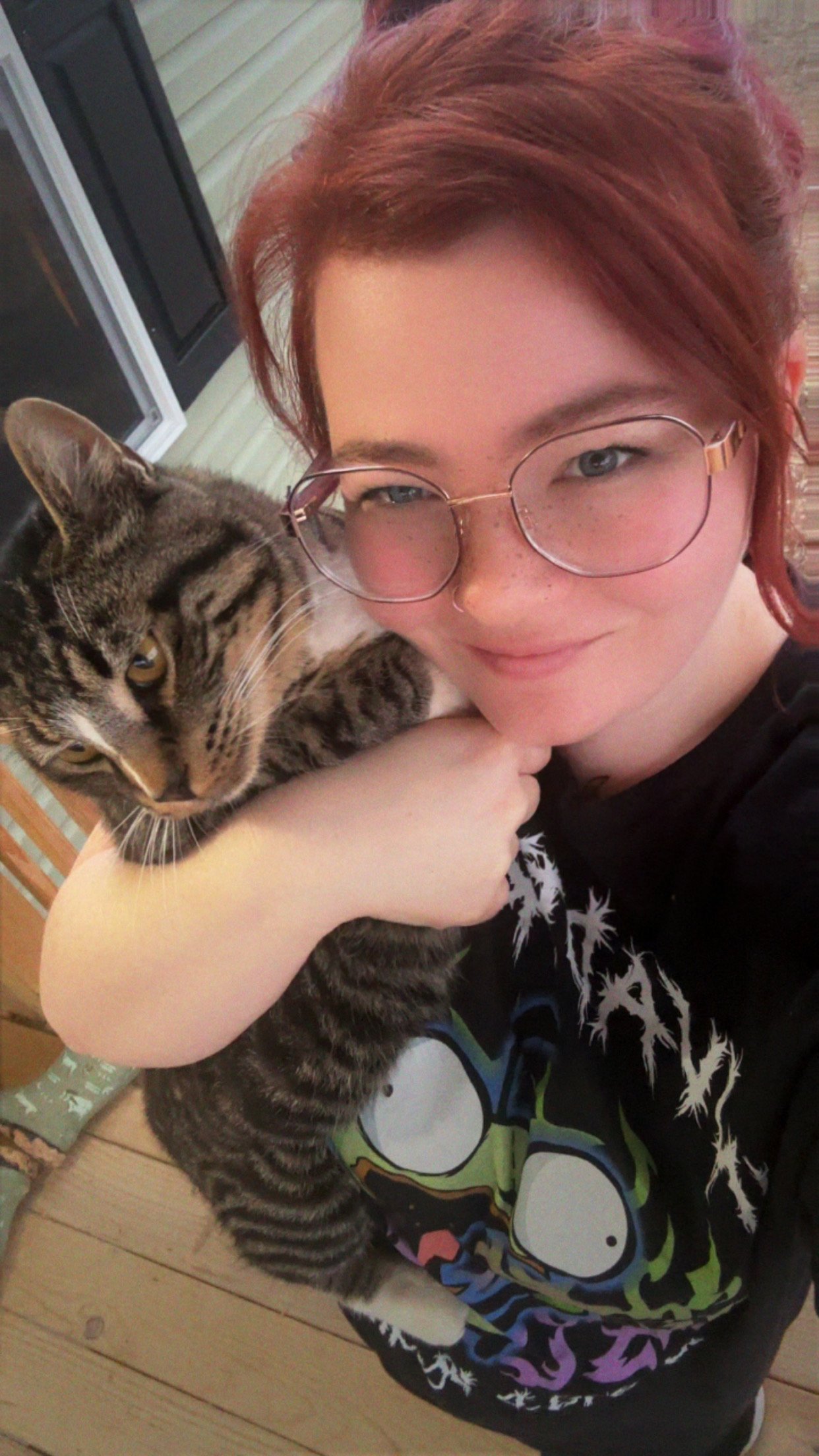 A young woman with red hair and glasses holds a tabby cat close to her face, both looking into the camera on a wooden porch.