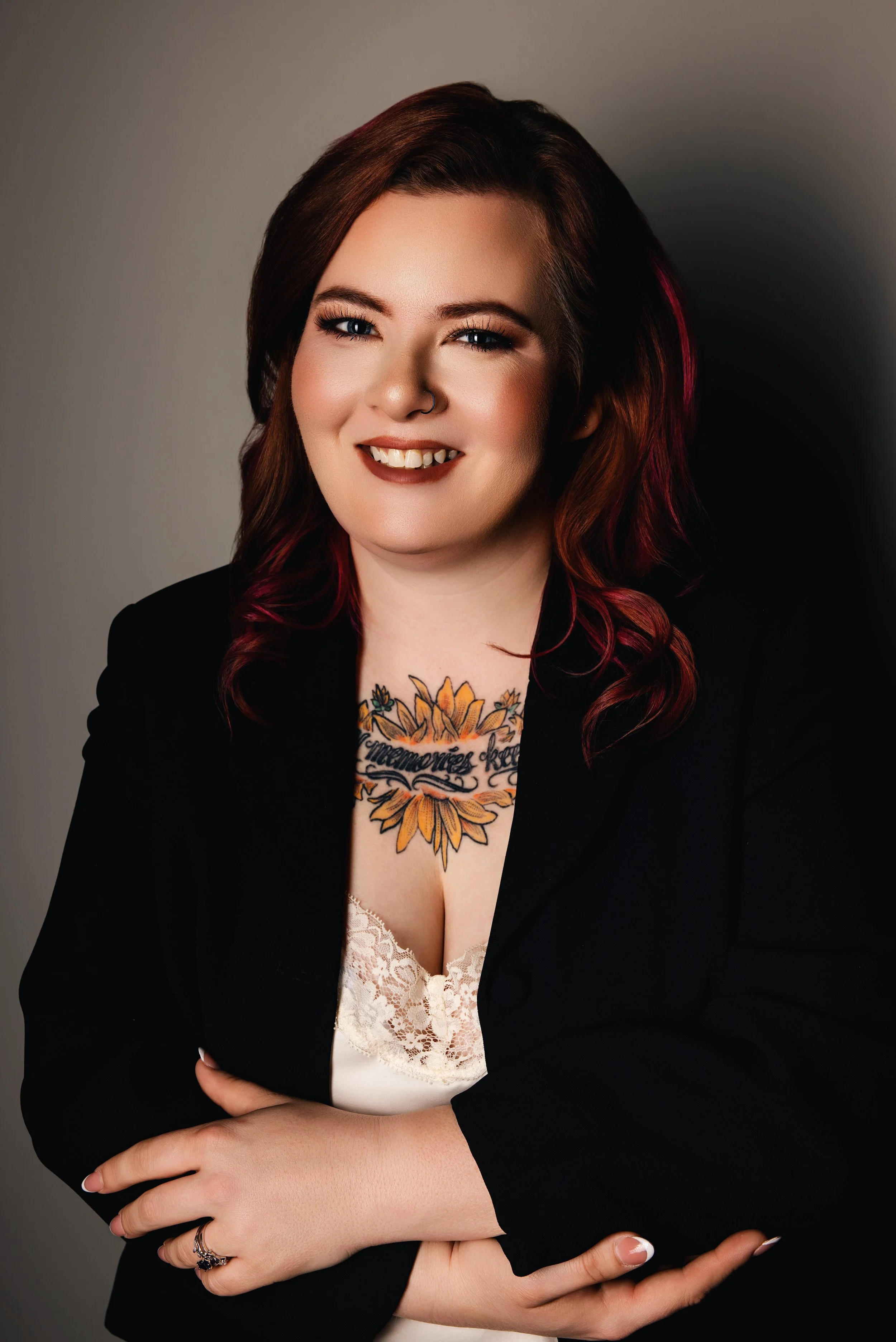 A woman with auburn hair styled in loose waves, wearing a black blazer over a lace-trimmed top, is smiling and looking at the camera, with a tattoo of a sunflower and text on her chest.