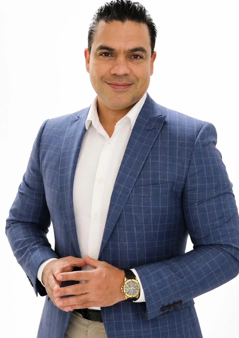 A man in a blue checkered suit with a white shirt, standing with his hands clasped, wearing a gold watch, smiling against a plain white background.