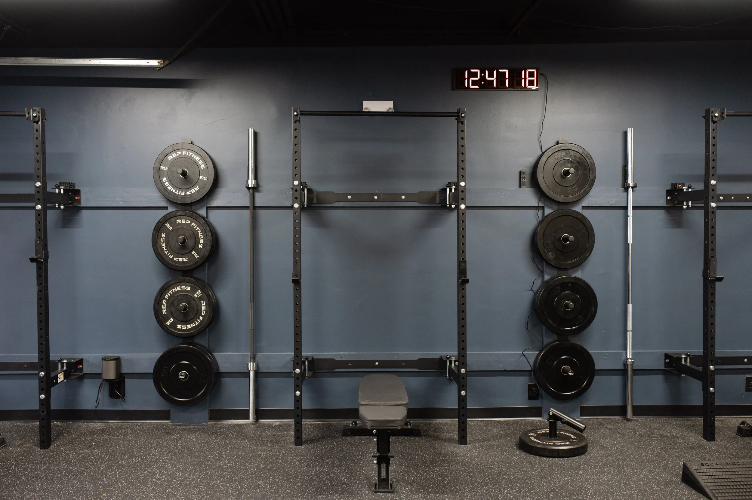 Empty gym workout area with weight plates on wall, black squat rack, small stool, and digital clock showing 12:47:18.
