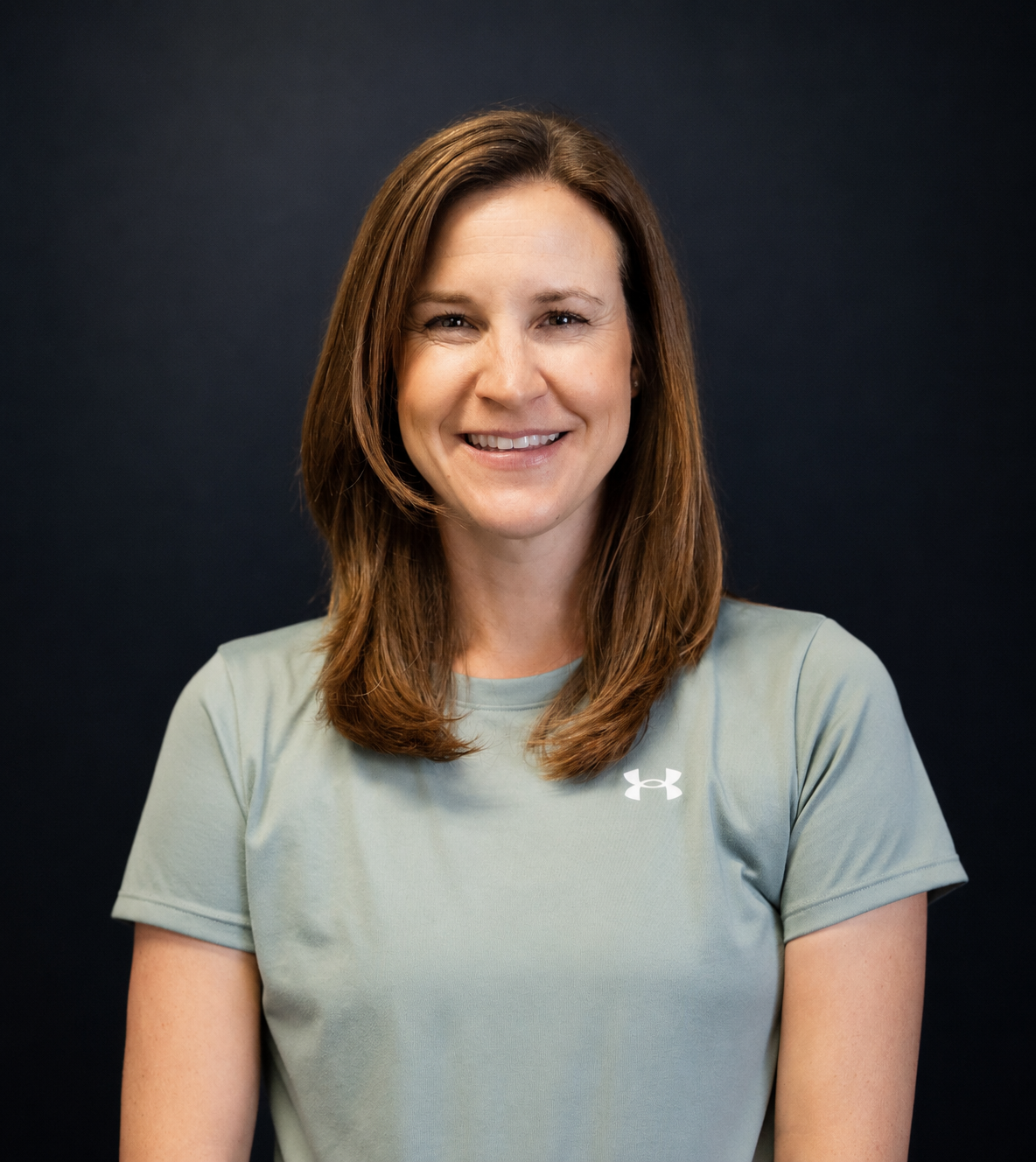 A woman with shoulder-length brown hair, smiling, wearing a light gray Under Armour t-shirt, against a dark background.