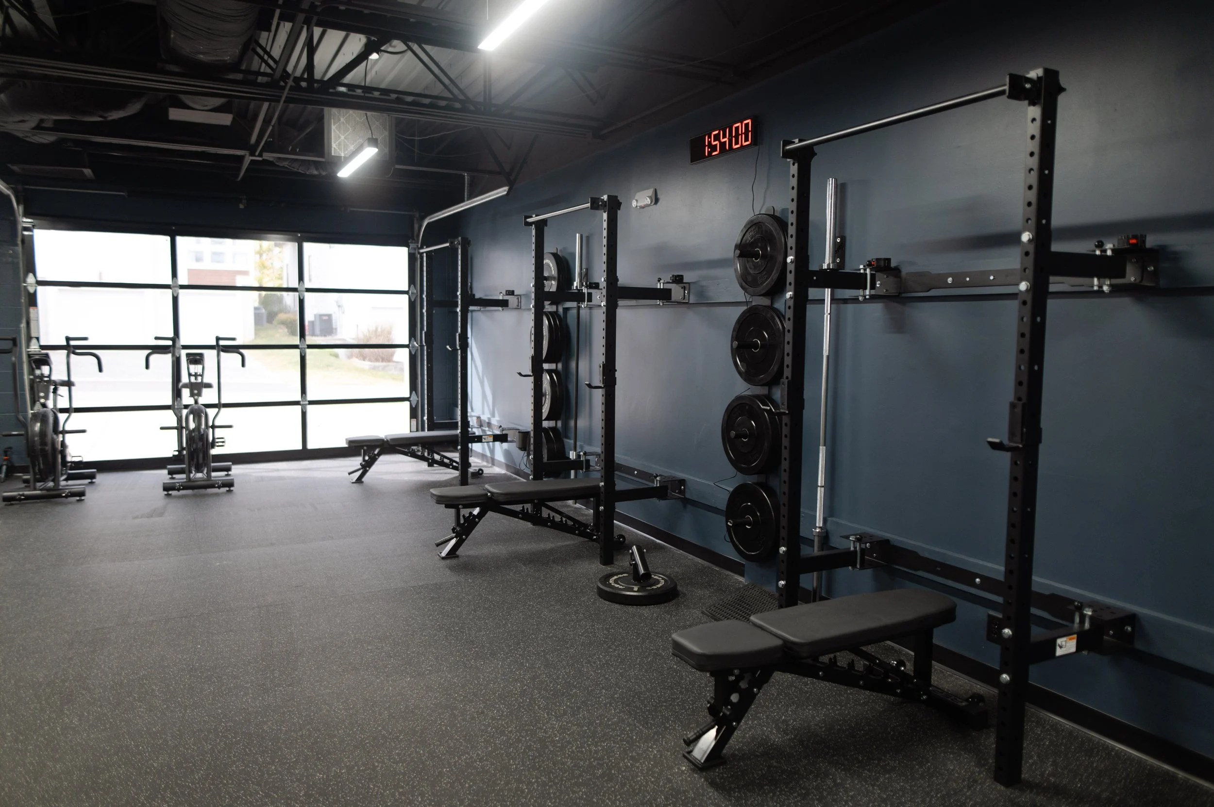 Interior of a modern gym with black metal workout equipment, including weight racks and benches, against a dark blue wall with a digital clock showing 1:54:00, large windows letting in natural light, and ceiling lights.