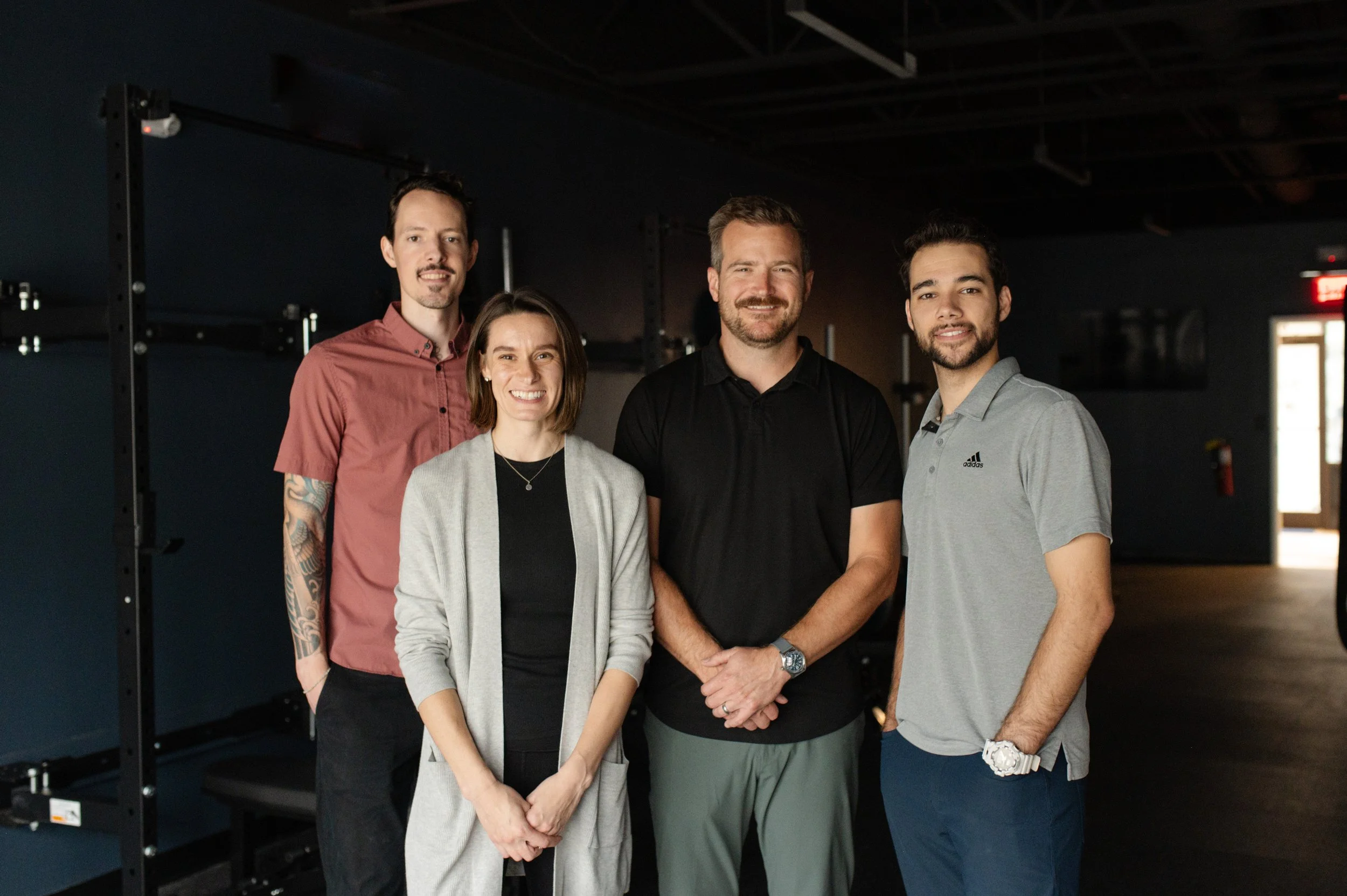 A group of four diverse adults smiling and standing together in a dimly lit indoor space, possibly a gym or fitness center.