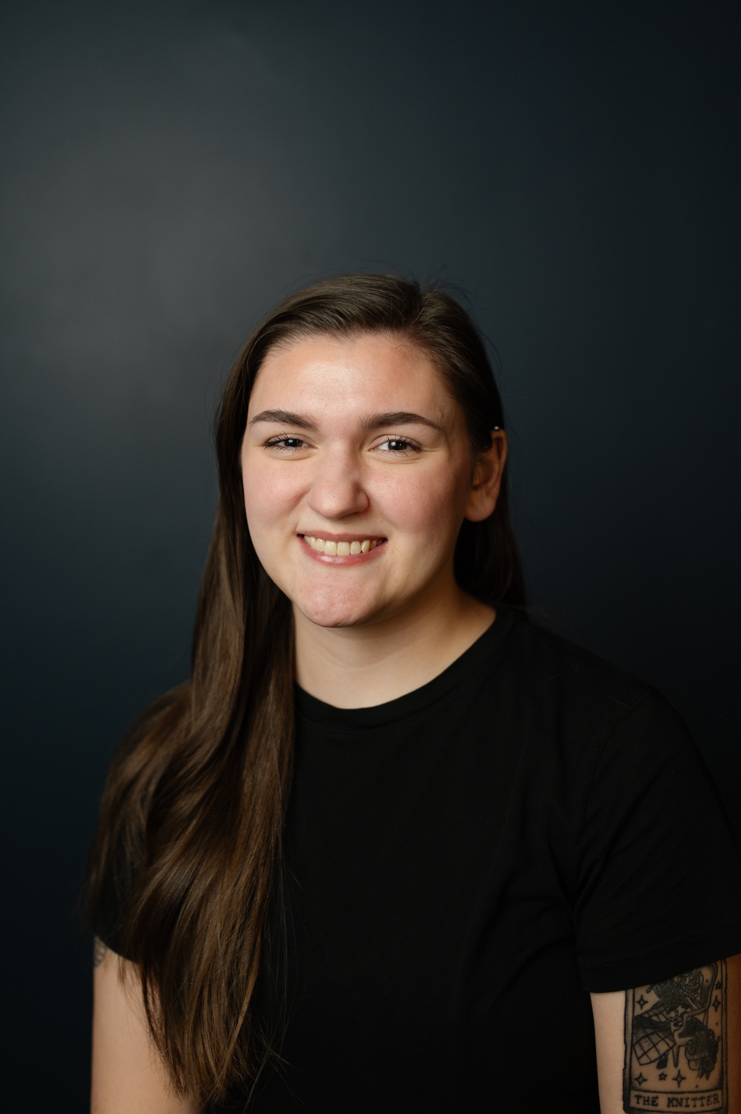 Portrait of a young woman with long brown hair, smiling, wearing a black shirt, against a dark background.