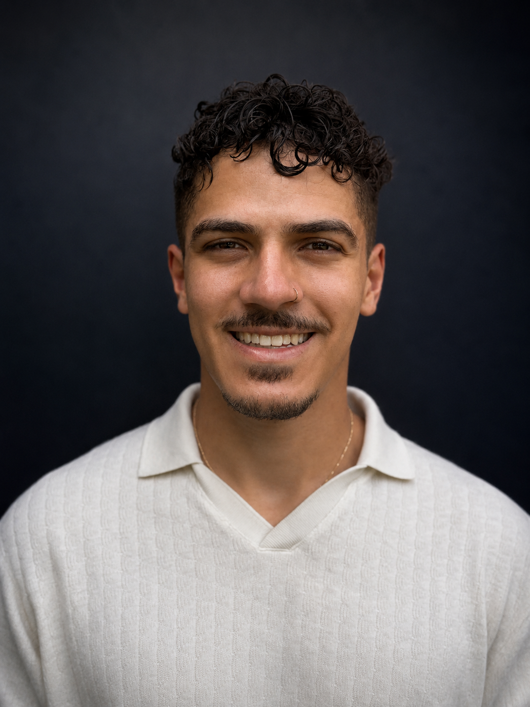 A young man with curly dark hair, a nose piercing, and a goatee, smiling in front of a dark background, wearing a cream-colored sweater.