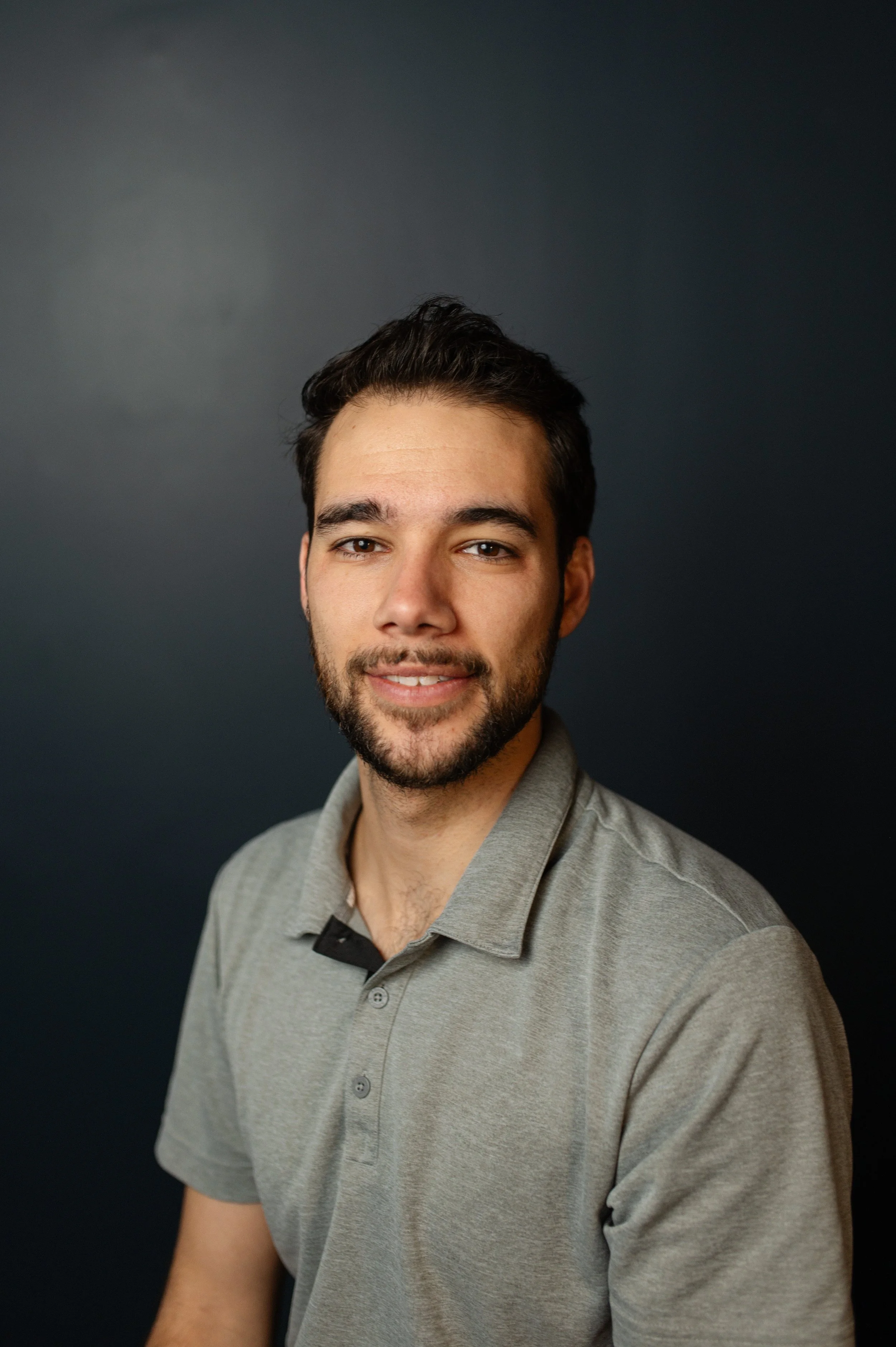 A smiling young man with dark hair, beard, and mustache, wearing a gray polo shirt, standing against a dark background.