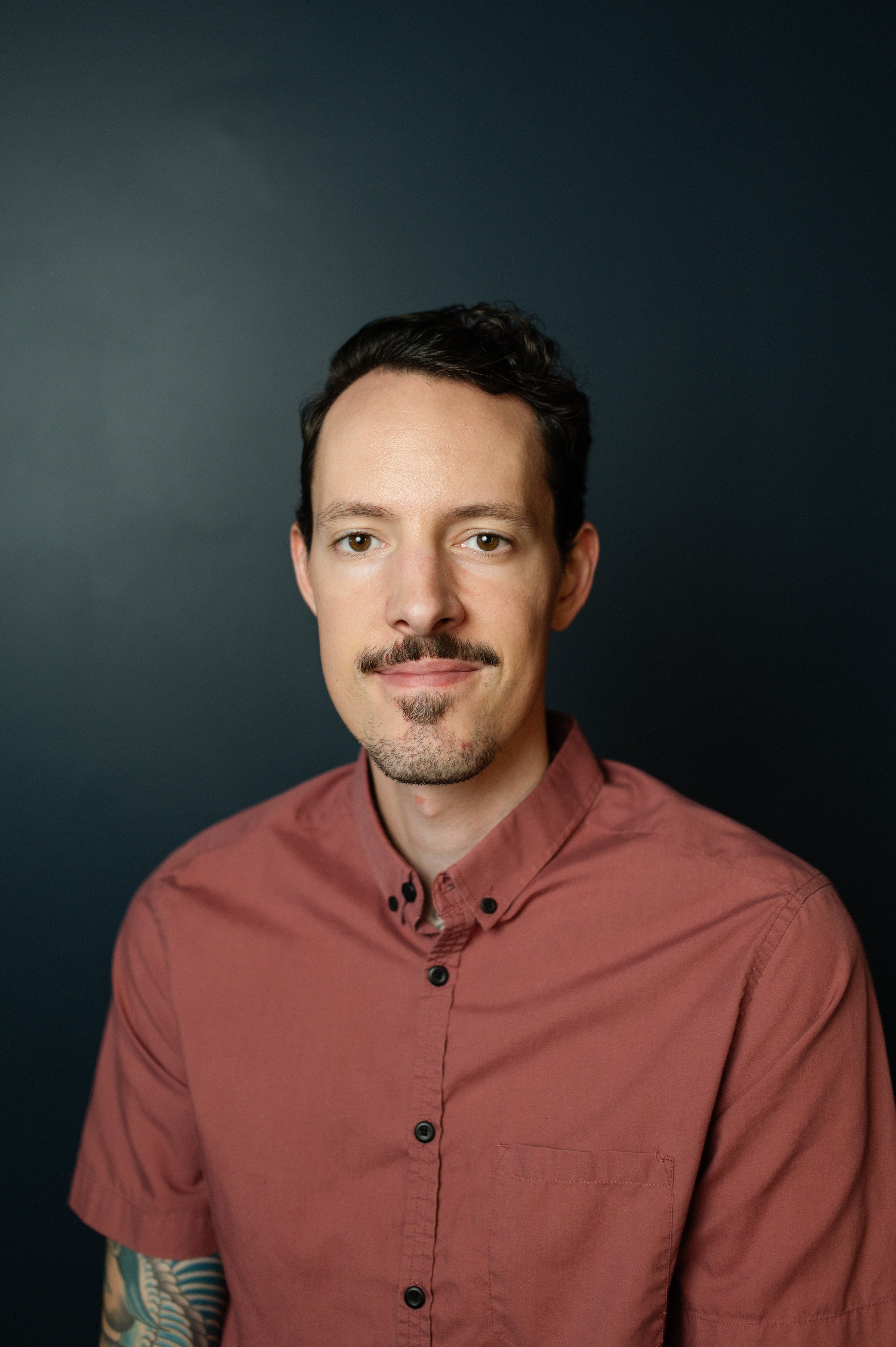 A portrait of a man with short dark hair, a mustache, and a goatee, smiling slightly, wearing a rust-colored button-up shirt against a dark background.