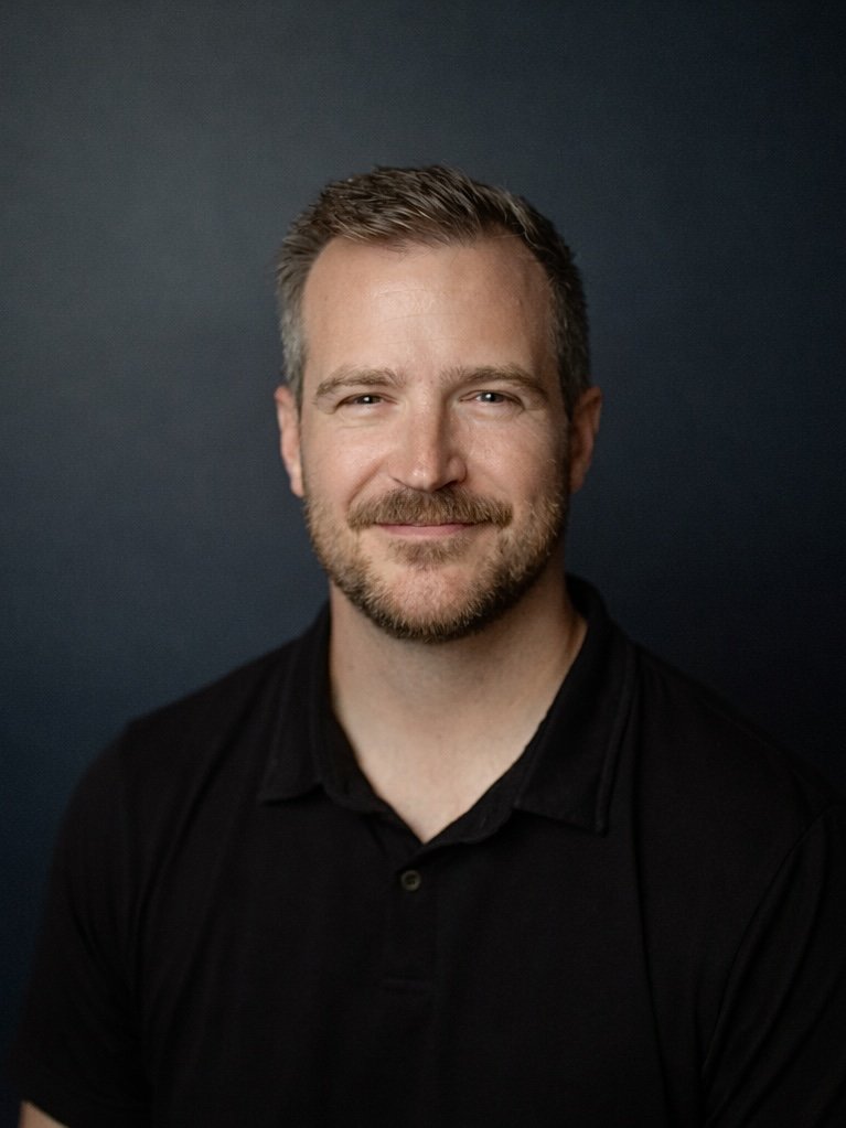 A man with short brown hair, a beard, and wearing a black shirt, smiling at the camera against a dark background.