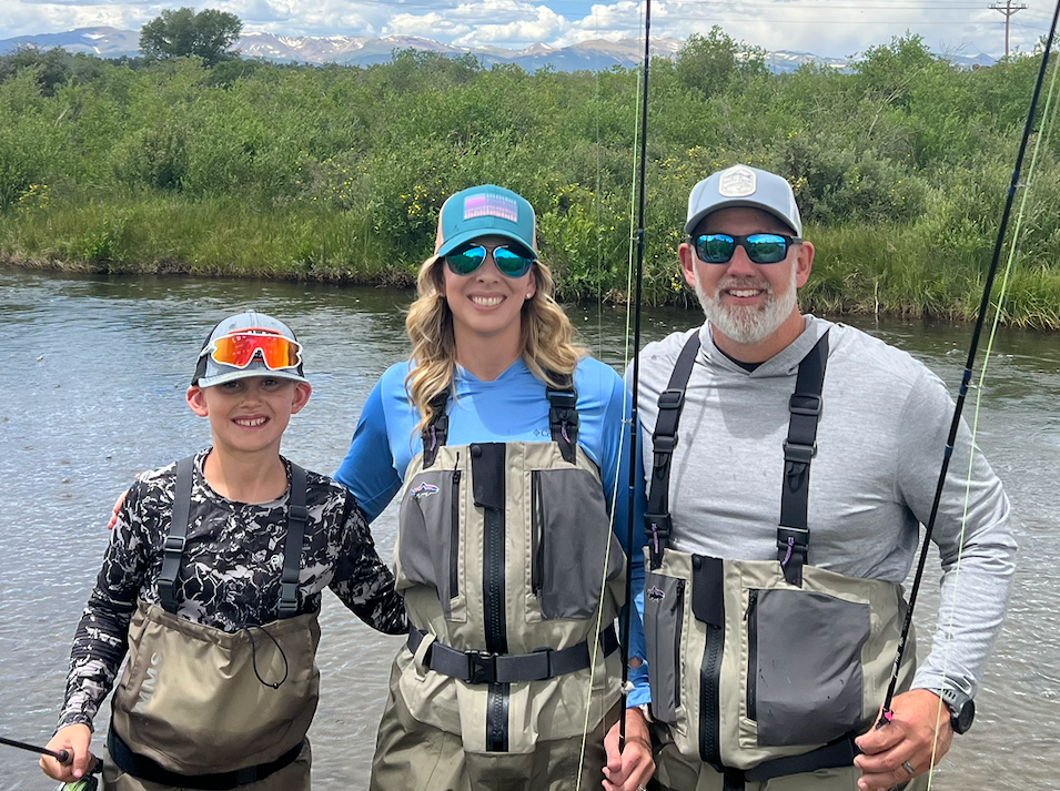 Family fishing by a river with green bushes and mountains in the background, all wearing fishing gear and sunglasses.