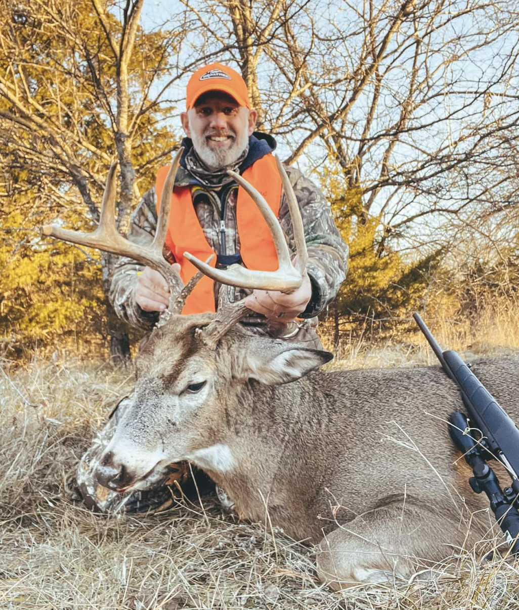 A man in camouflage clothing and an orange safety vest holds the antlers of a large deer with brown fur and white markings, lying on the grass in a wooded area during autumn.
