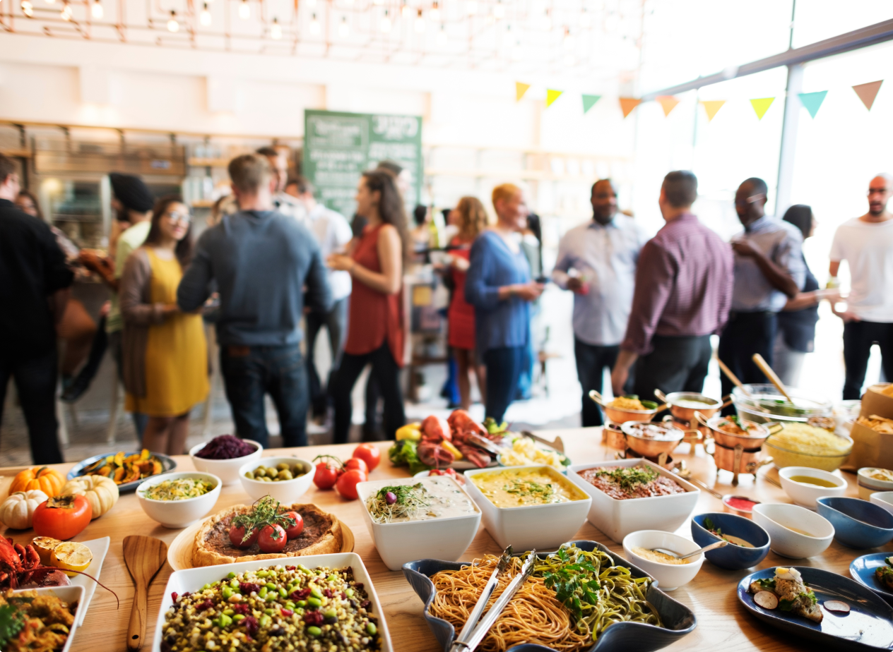 Buffet table with salads, pasta, and vegetables at a social gathering with people talking in a bright indoor space decorated for an event.