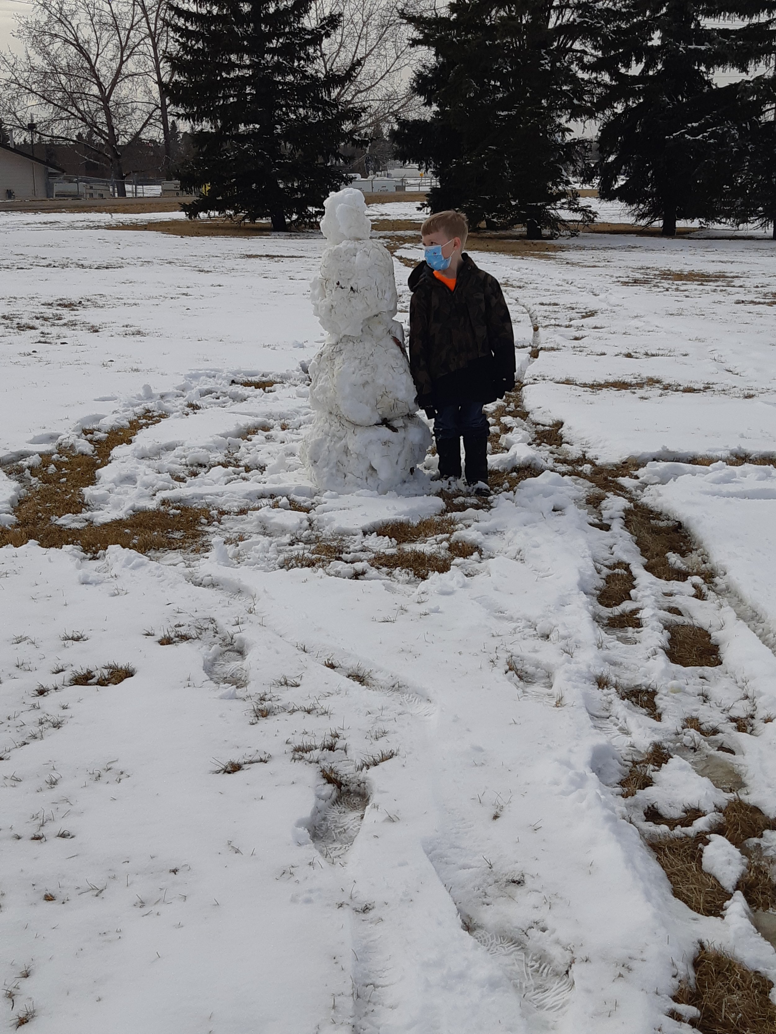 A boy wearing a face mask and black jacket stands outside in a snow-covered field next to a small snowman. The snowman is built with snowballs and has a smaller snowball on top for a hat. There are tire tracks and footprints in the snow, with patches of brown grass visible.