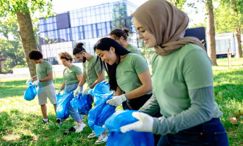 Group of diverse volunteers cleaning a park, holding blue trash bags and wearing gloves, with trees and a modern building in the background.