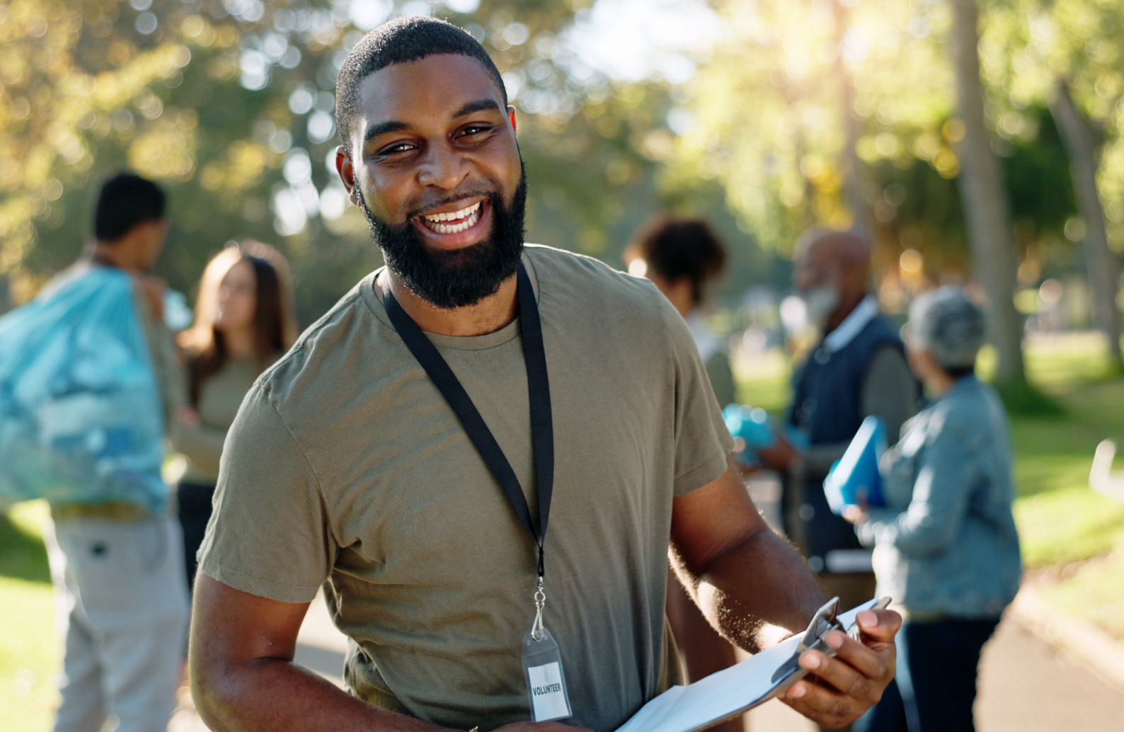 A smiling man with a beard wearing a volunteer badge, holding a clipboard, standing outdoors in a park with other people in the background.