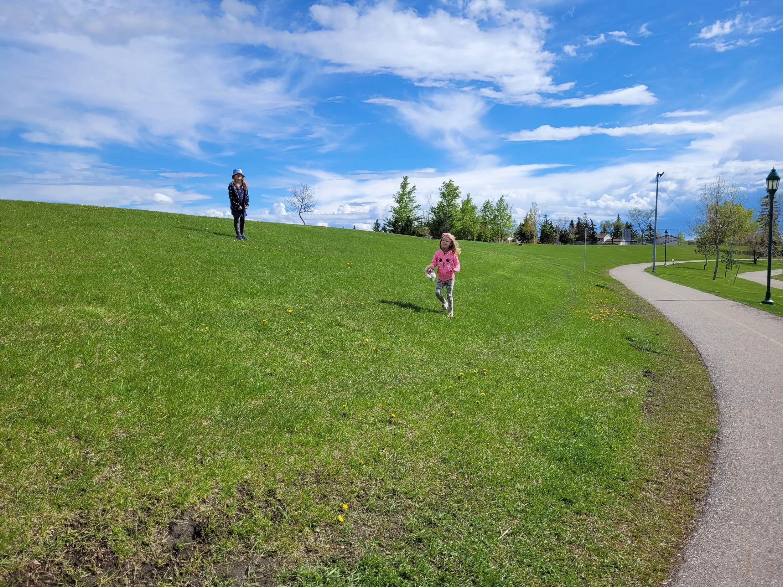 Two young girls playing on a grassy hill underneath a partly cloudy blue sky. One girl wears a pink hoodie and grey pants, the other girl wears a hat, dark jacket, and patterned leggings. There are trees, a pathway, and street lamps visible in the background.