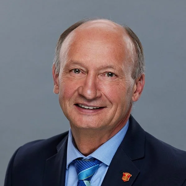 A middle-aged man with a friendly expression wearing a dark suit, light blue shirt, and a blue striped tie, standing against a gray background.