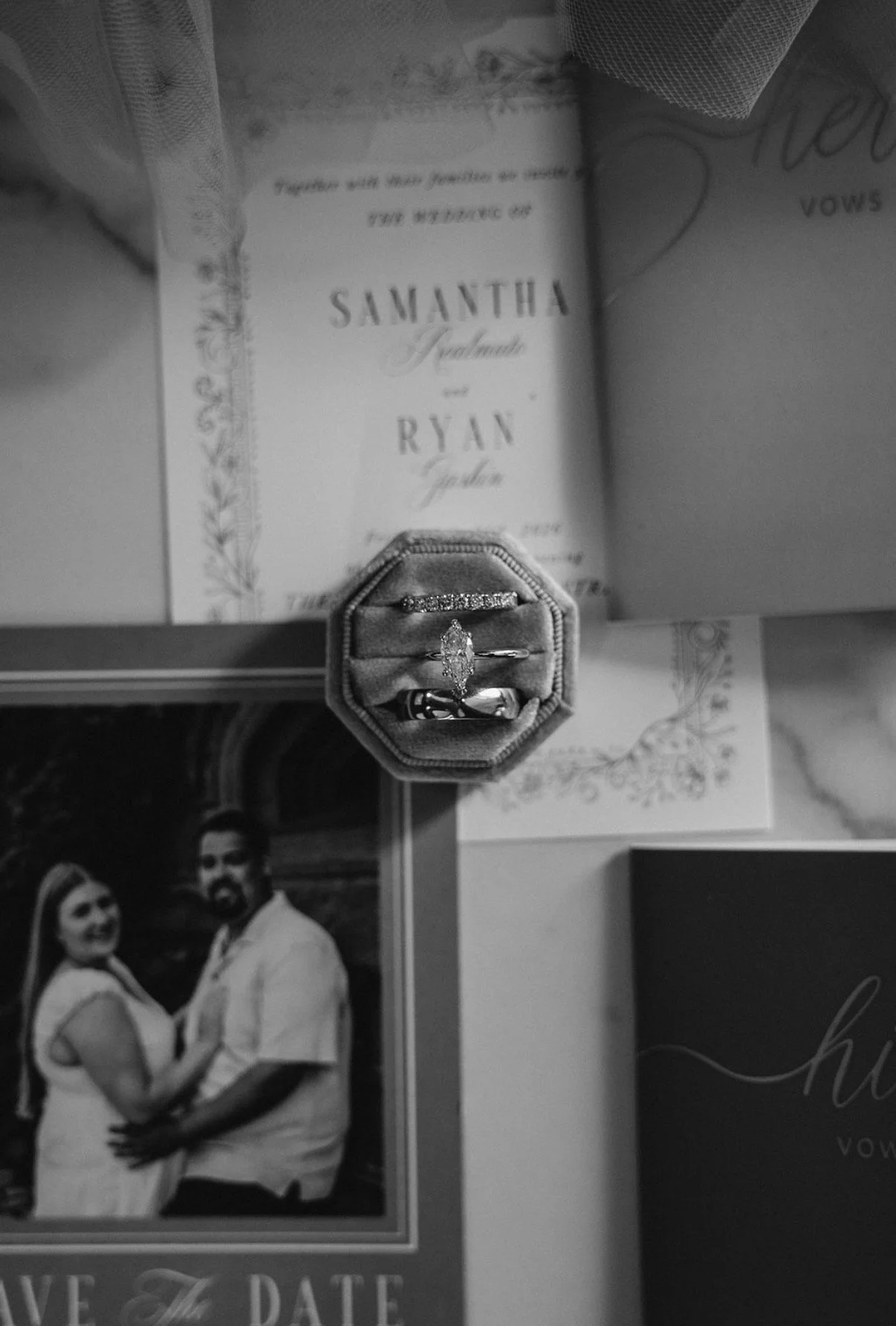 Black and white photo of a wedding ring set in a small jewelry box, placed on a table with wedding invitations and a photo of a couple in the background.