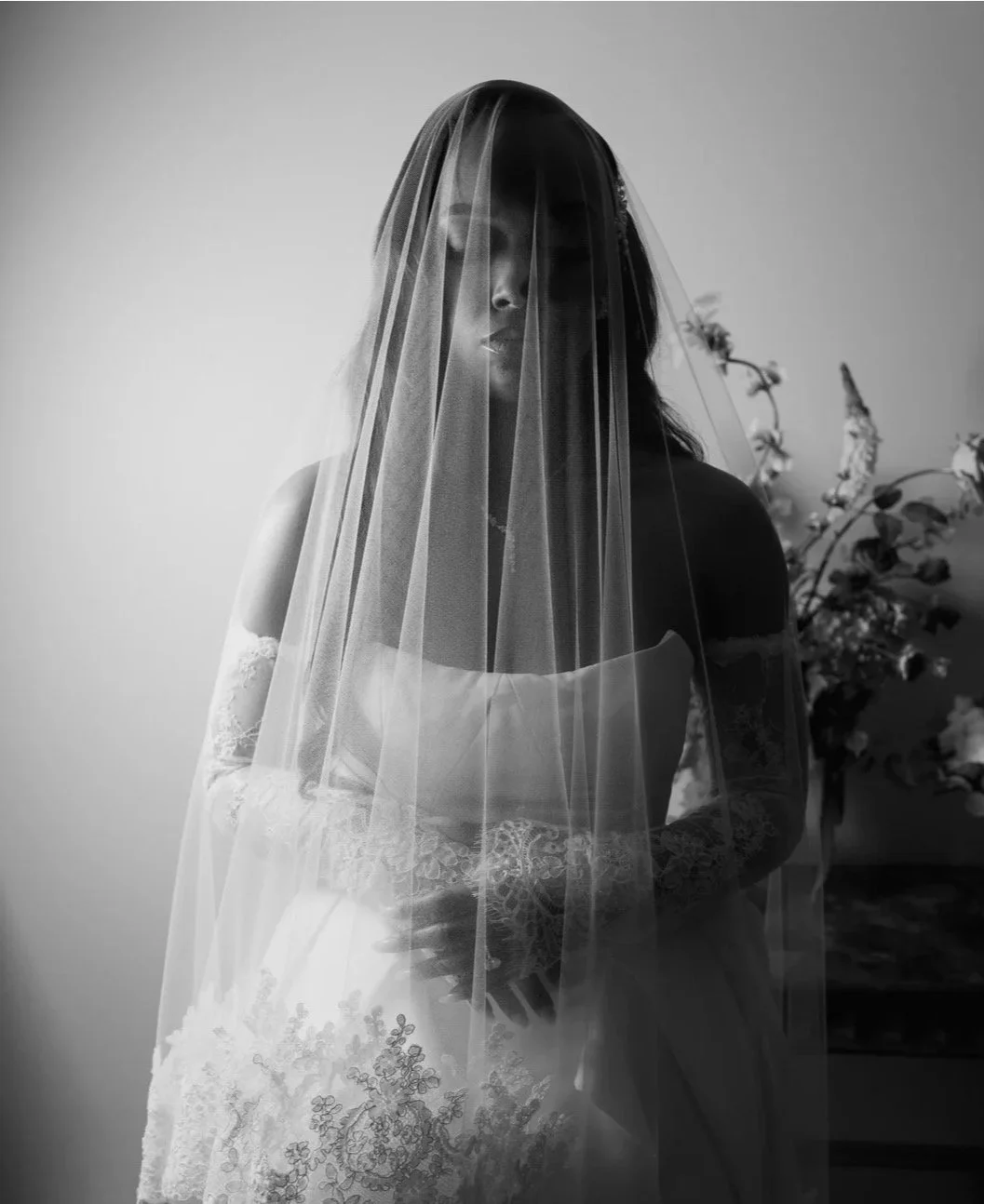A black and white photo of a woman dressed in a wedding gown with lace details, wearing a veil that covers her face and head, standing in front of a plain wall and some flowers in the background.