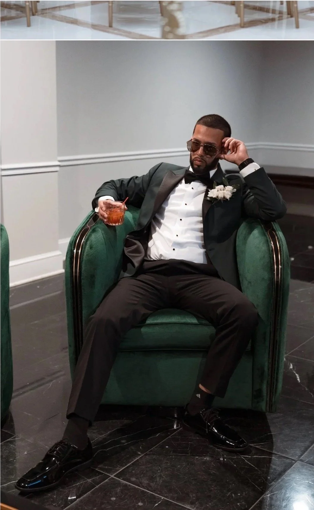 A man in a tuxedo sitting in a green velvet armchair, holding a drink, wearing sunglasses, and relaxing with one arm resting on the armrest and the other adjusting his sunglasses.
