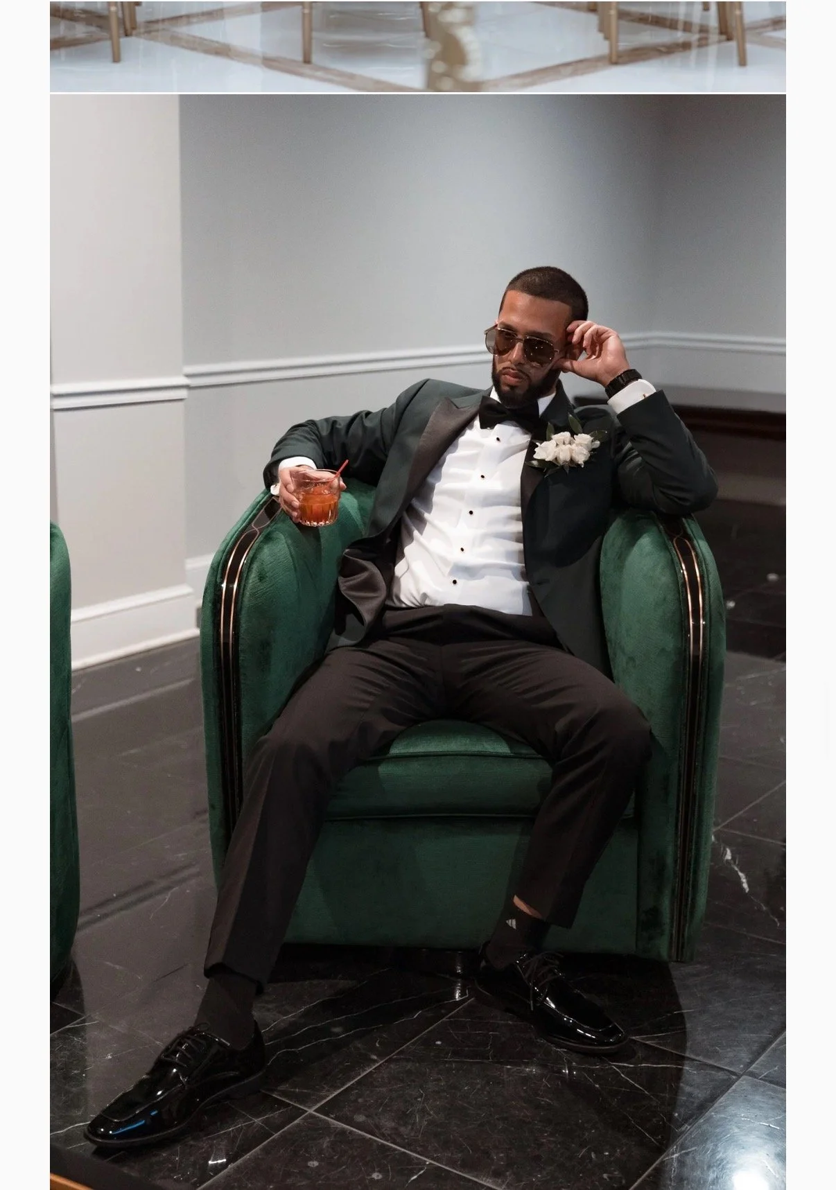 A man in a tuxedo sitting in a green velvet armchair, holding a drink, wearing sunglasses, and relaxing with one arm resting on the armrest and the other adjusting his sunglasses.