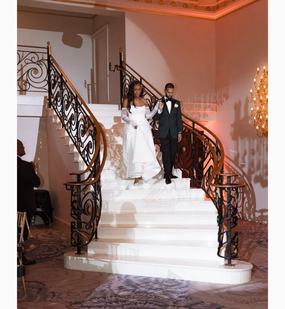 A bride and groom descending a grand staircase at a wedding. The bride is wearing a white wedding gown with lace sleeves, and the groom is in a black tuxedo with a bow tie and boutonniere. They are holding hands, surrounded by elegant decor including a chandelier and ornate staircase railing.