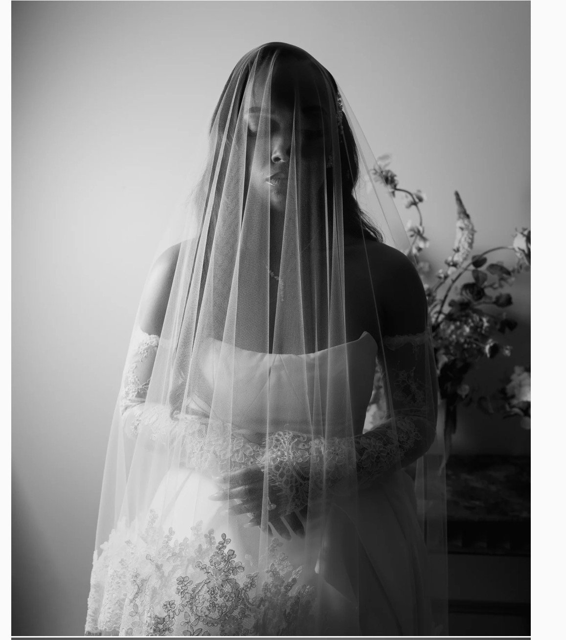 A black and white photo of a woman dressed in a wedding gown with lace details, wearing a veil that covers her face and head, standing in front of a plain wall and some flowers in the background.
