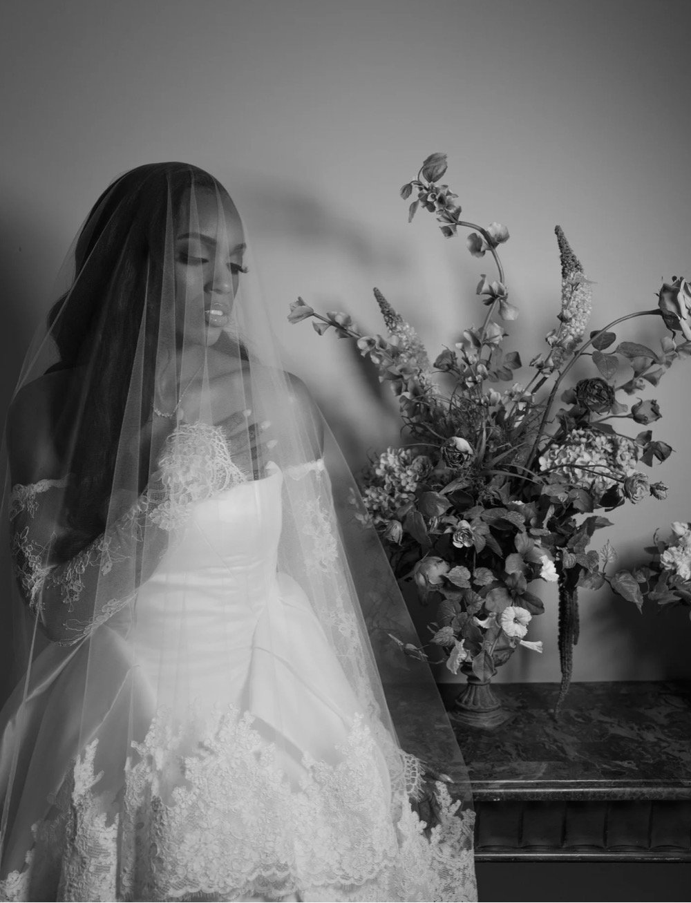 A wedding dress mannequin with a veil and lace details, positioned next to a large floral arrangement with various flowers and greenery on a table.