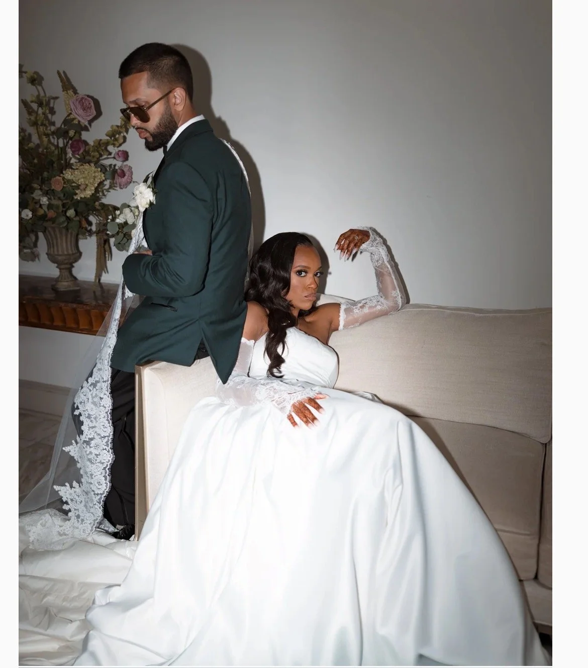 A woman in a white wedding dress and lace gloves sitting on a couch with henna on her hands, and a man in a suit with sunglasses standing beside her, in an indoor setting with a floral arrangement in the background.
