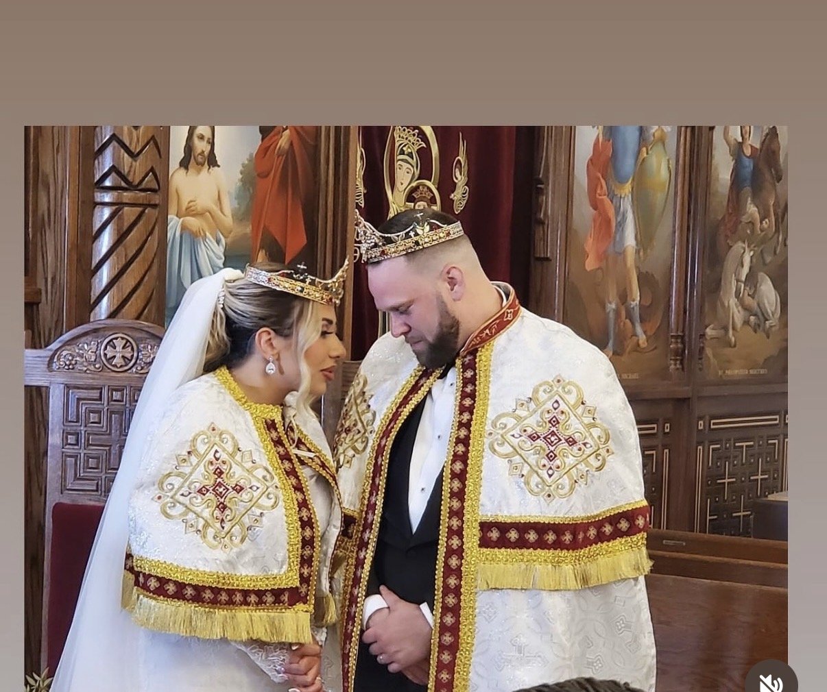 Bride and groom wearing crowns and ceremonial cloaks during their wedding in a church, holding hands with heads bowed.