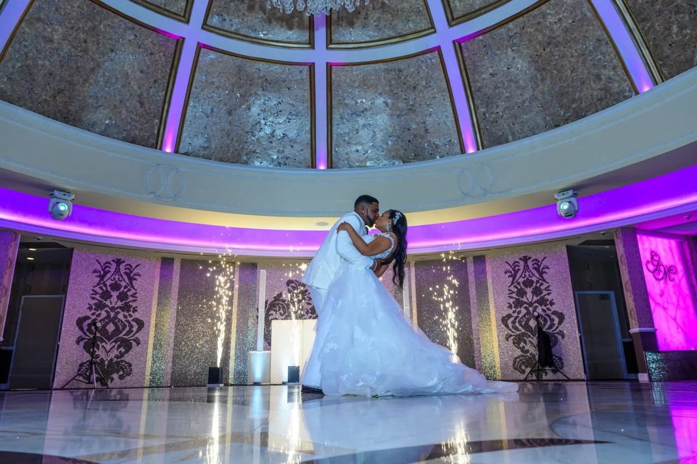 A bride and groom are dancing together in a ballroom decorated with purple and pink lighting, with sparklers on the floor behind them.