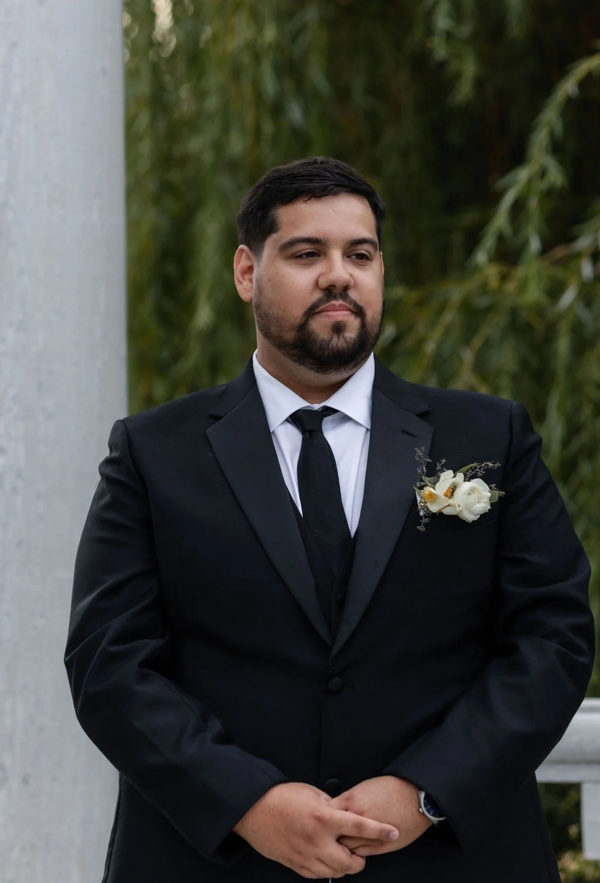 Man in black suit with a white shirt and black tie, standing outdoors with greenery in the background, wearing a white floral boutonniere on his lapel.