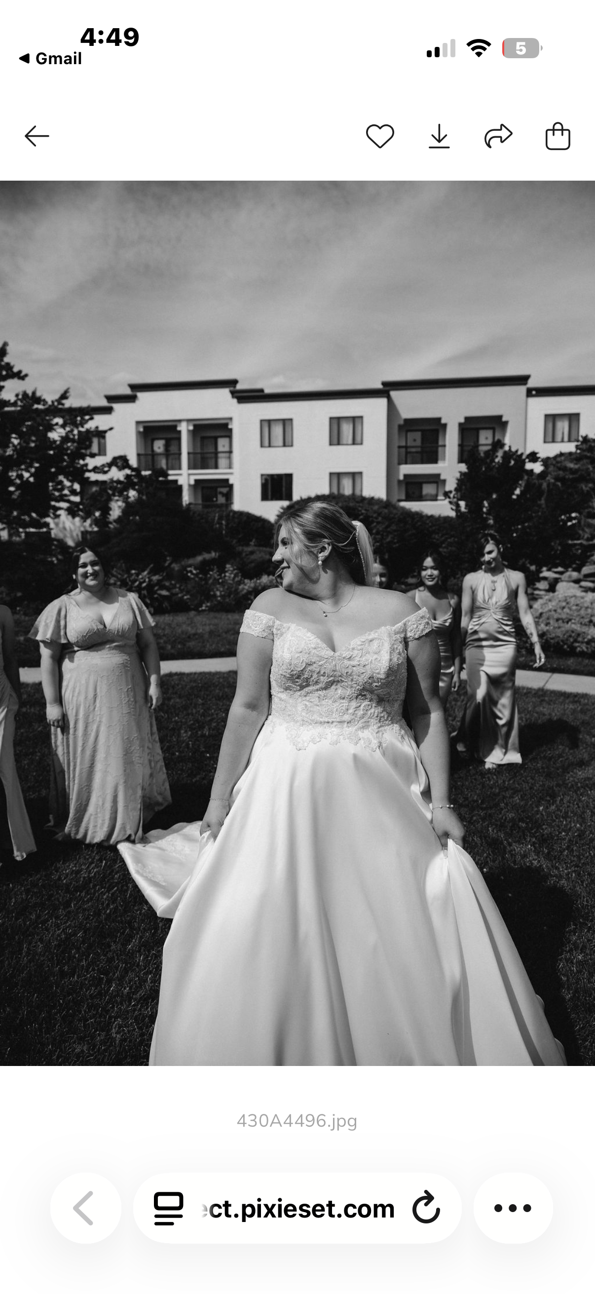 A group of women, including a bride in a wedding gown, standing on a grassy area outside a building with multiple balconies. The bride is in the foreground, smiling and holding her dress, with her hair in a ponytail. The other women are dressed in fo
