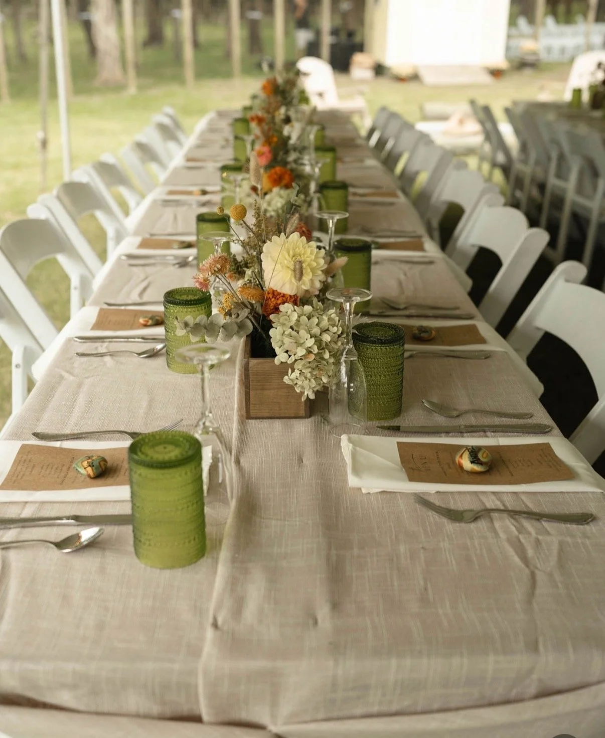 Long outdoor dining table decorated with floral centerpiece, green candles, glassware, and place settings with paper menus and eggs with colorful designs, set in a garden or park setting.