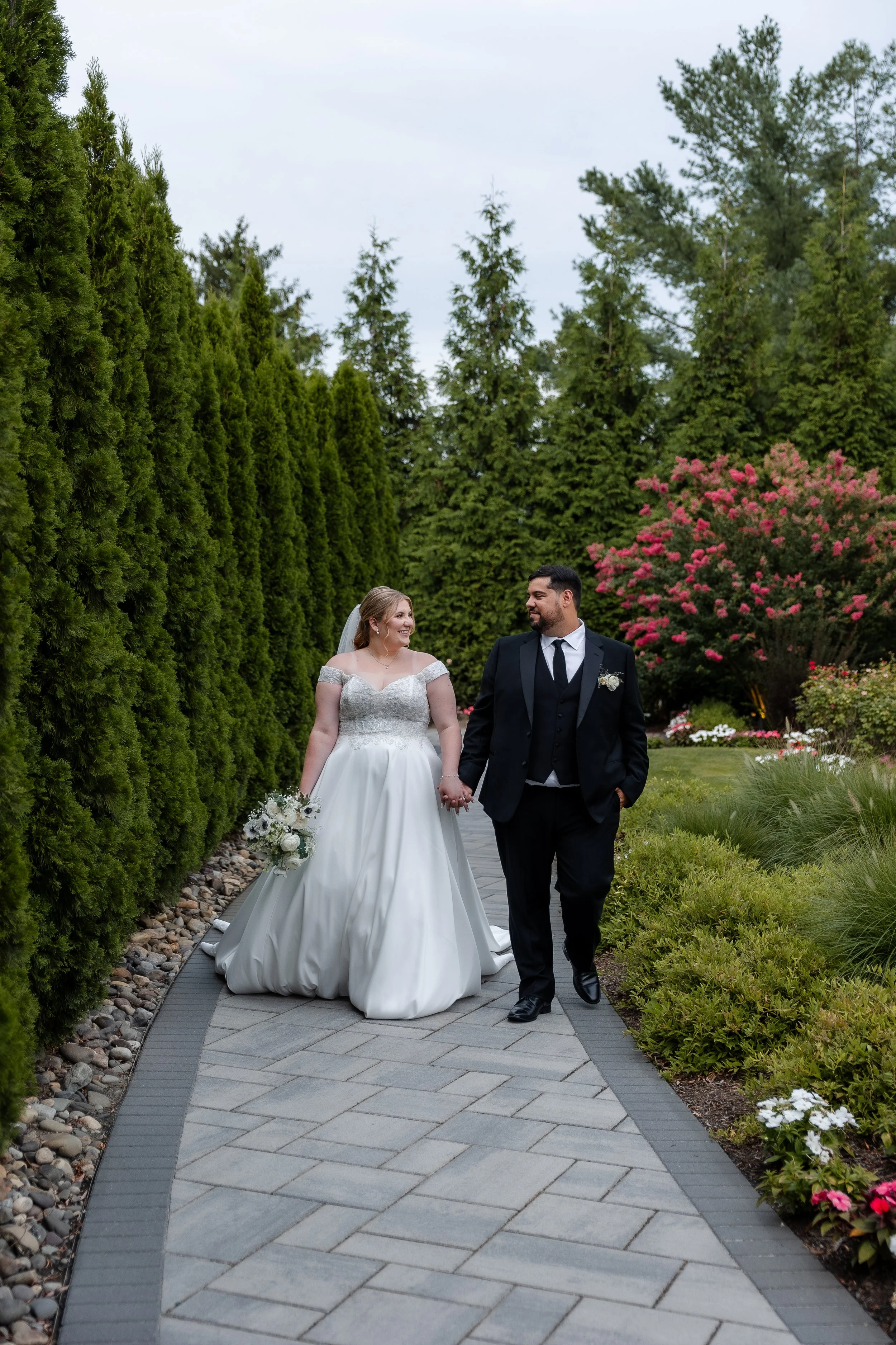 A bride and groom walk hand in hand down a garden pathway, surrounded by lush green trees and colorful flowering bushes on a cloudy day.