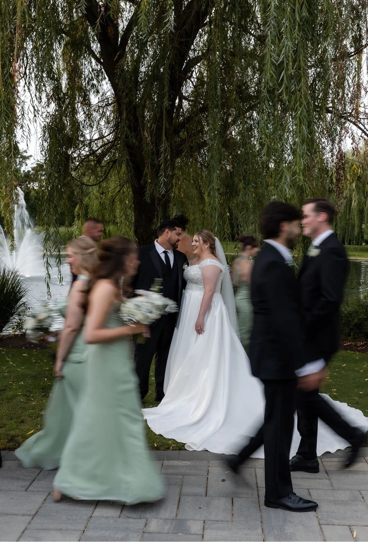 A bride and groom stand under a large tree near a pond, smiling at each other during their wedding. Several wedding guests walk by in the foreground, some blurred due to motion. The setting is outdoors with greenery and a fountain in the background.