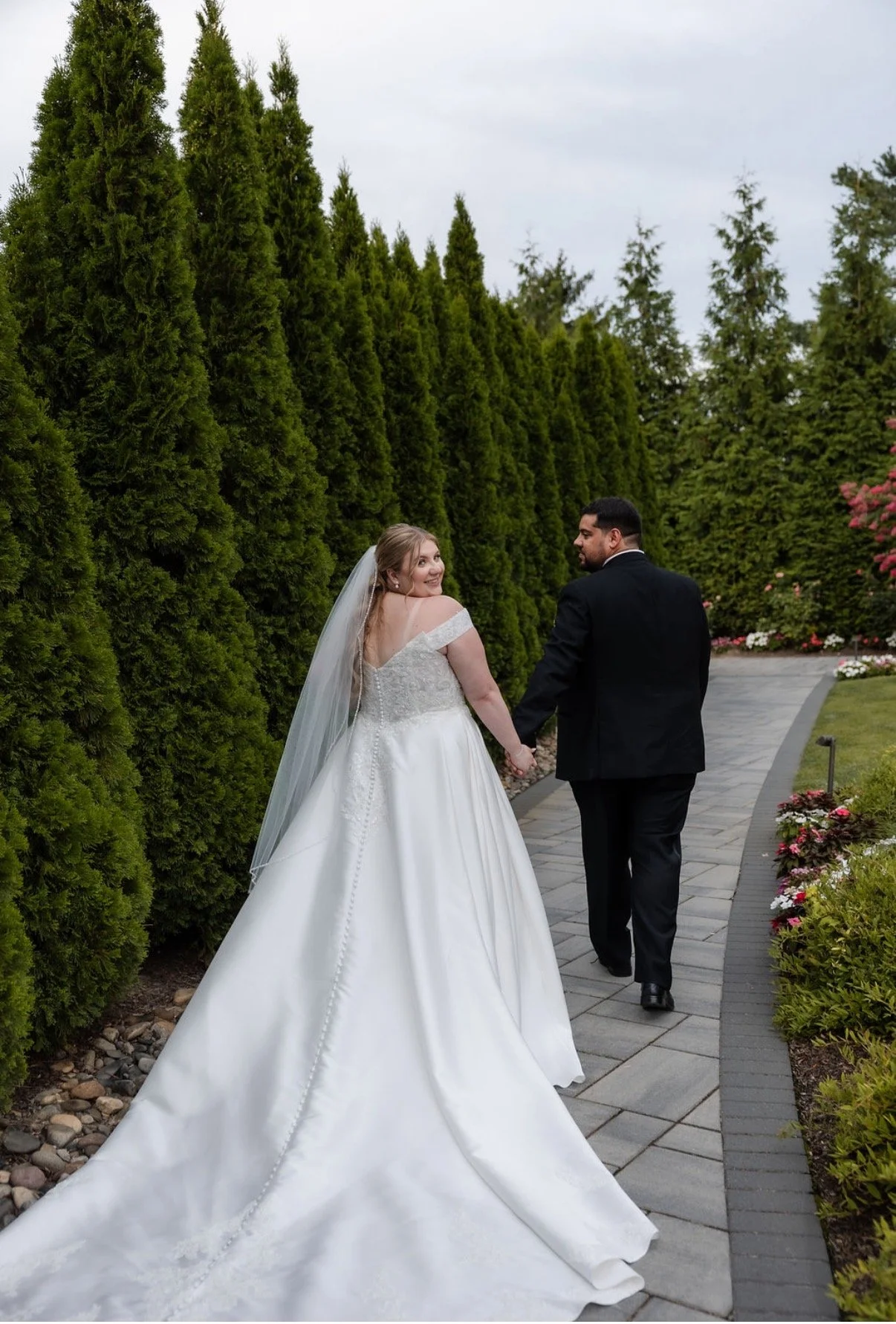 A bride and groom walk hand in hand outdoors on a garden pathway, surrounded by tall green trees and colorful flowers, with the bride smiling over her shoulder at the camera.