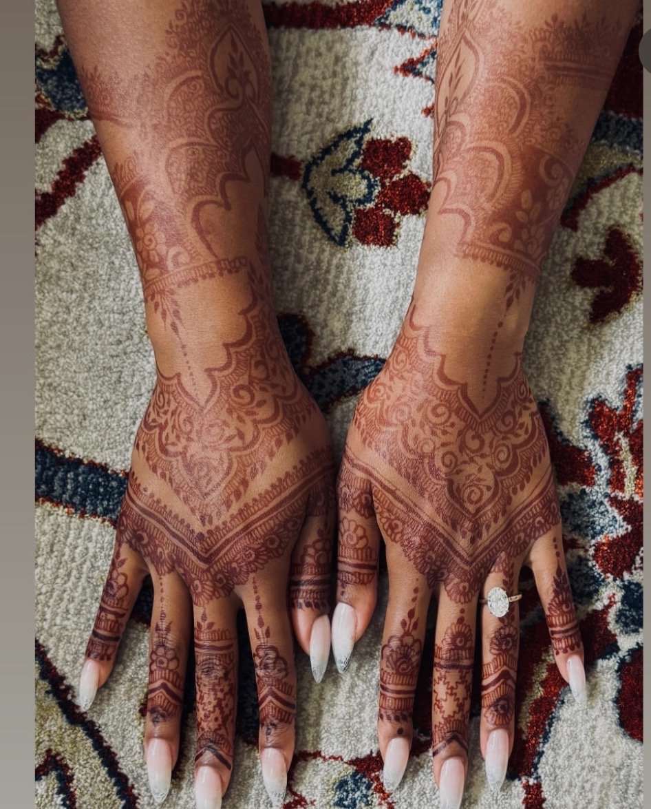 Tattooed hands with intricate henna designs resting on a patterned carpet.