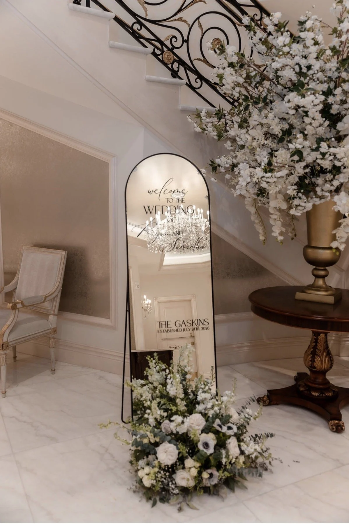 Elegant indoor wedding decoration with a large floral arrangement of white flowers and greenery, a decorative mirror with handwritten wedding welcome message, and a vintage-style chair and table in a luxurious setting.