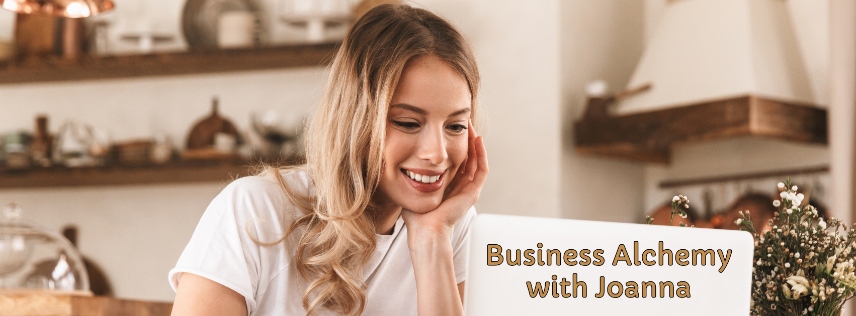A woman with long blonde hair smiling and looking at her laptop, which has the text "Business Alchemy with Joanna". The background shows a cozy, rustic kitchen with shelves and kitchenware.
