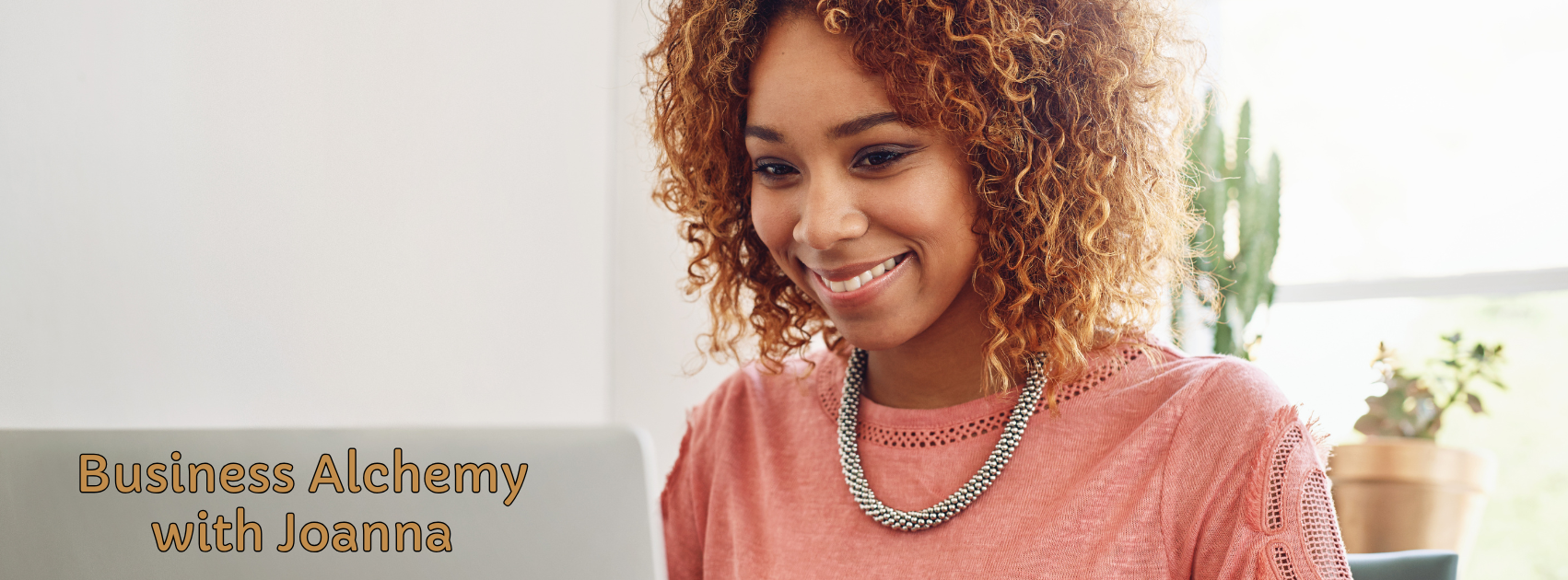 A woman with curly red hair smiling while working on a laptop. She is wearing a pink top and a beaded necklace. There are plants in the background.