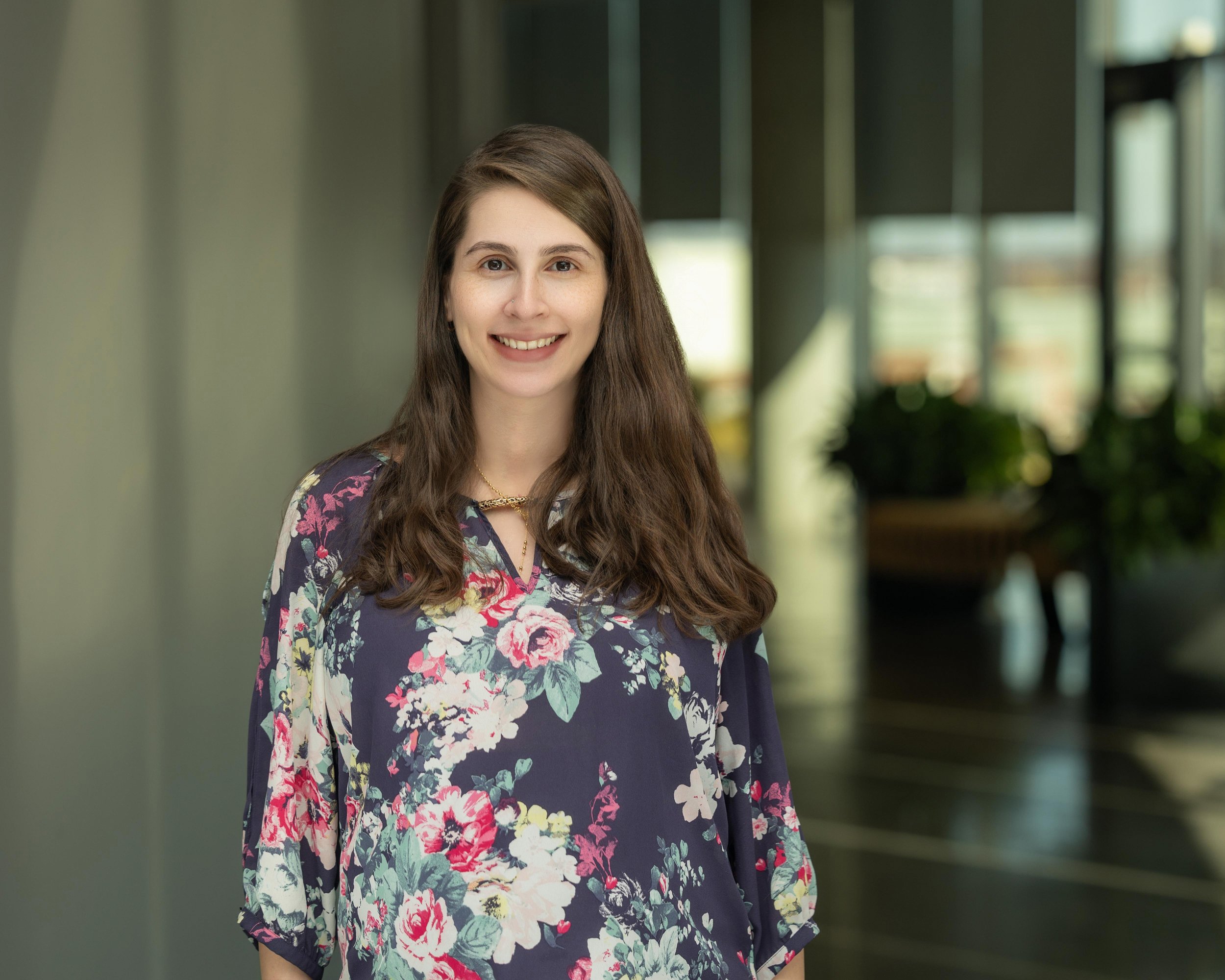 A professional woman with long brown hair, smiling, wearing a floral navy blue blouse, standing indoors with a blurred background of windows and indoor plants.