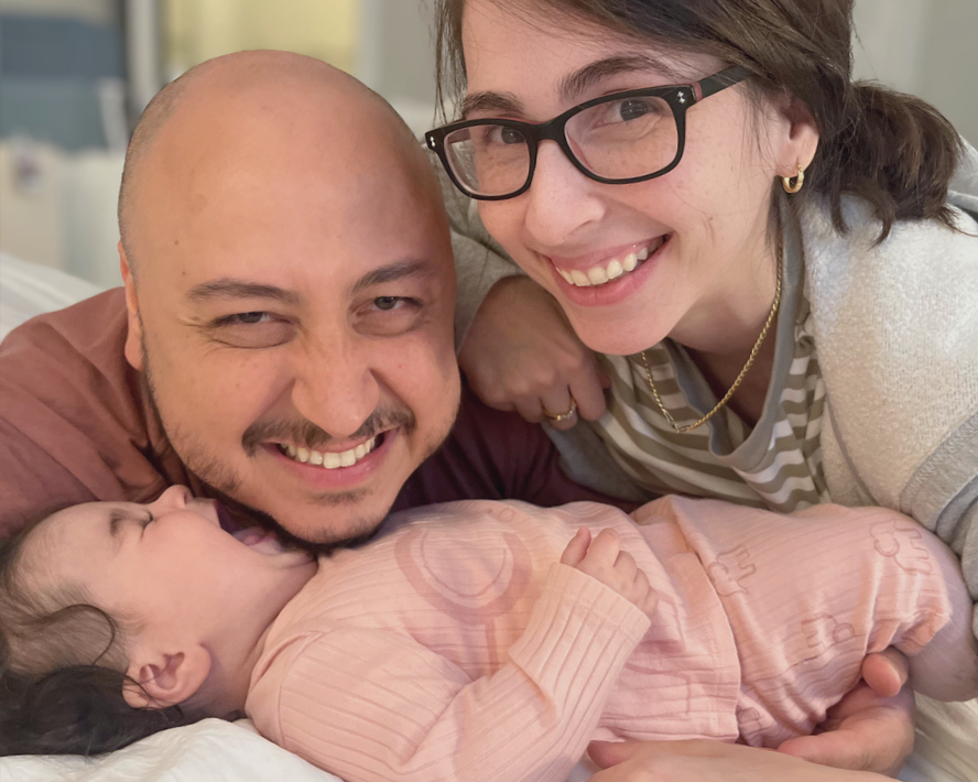 A happy family with a woman, a man, and a baby. The baby is lying on a bed, and the parents are smiling and leaning over the baby for a close-up shot.