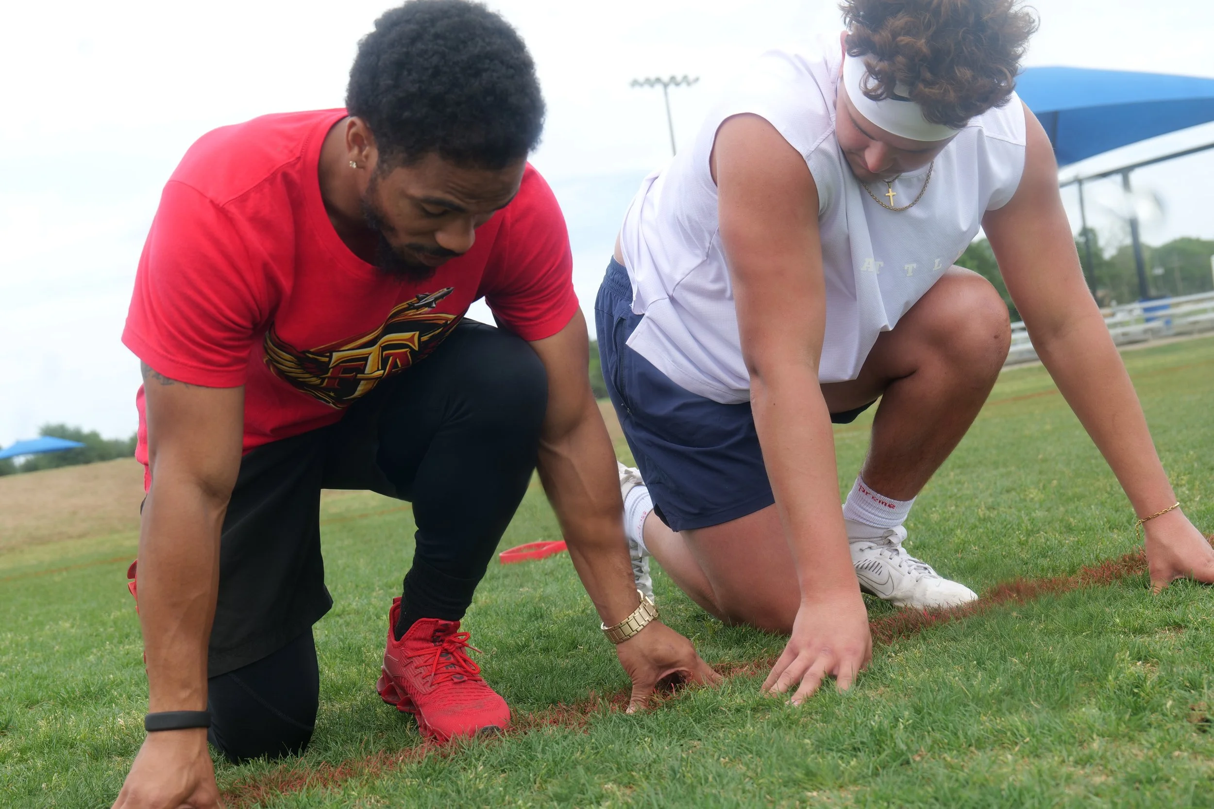 Two young men are doing push-ups on a grassy field, one in a red shirt and the other in a white shirt with a headband, near a sports field with bleachers and a blue shade structure in the background.