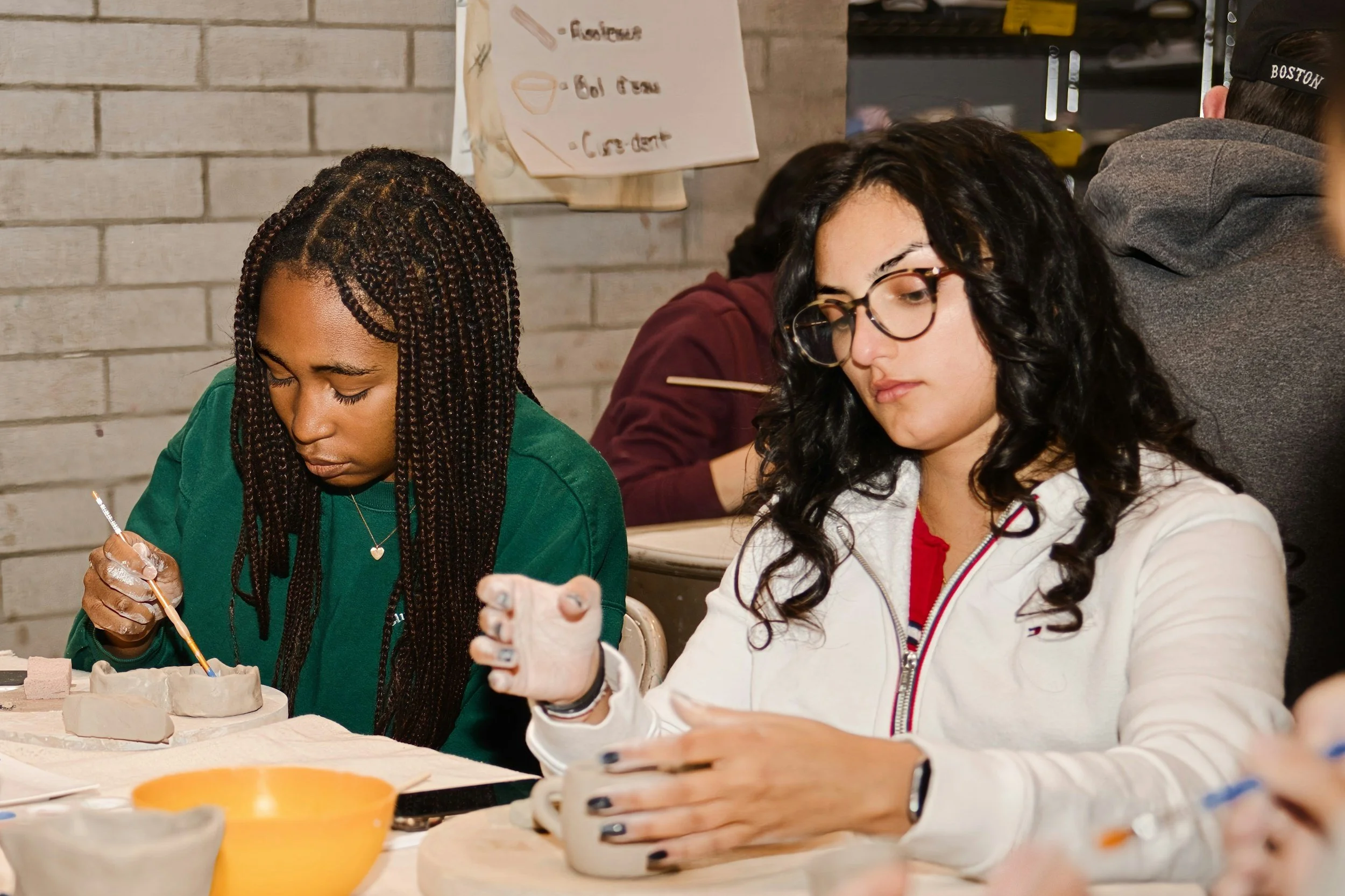 Two women working on a pottery project at a table, with a gray brick wall and a handwritten sign with instructions in the background.