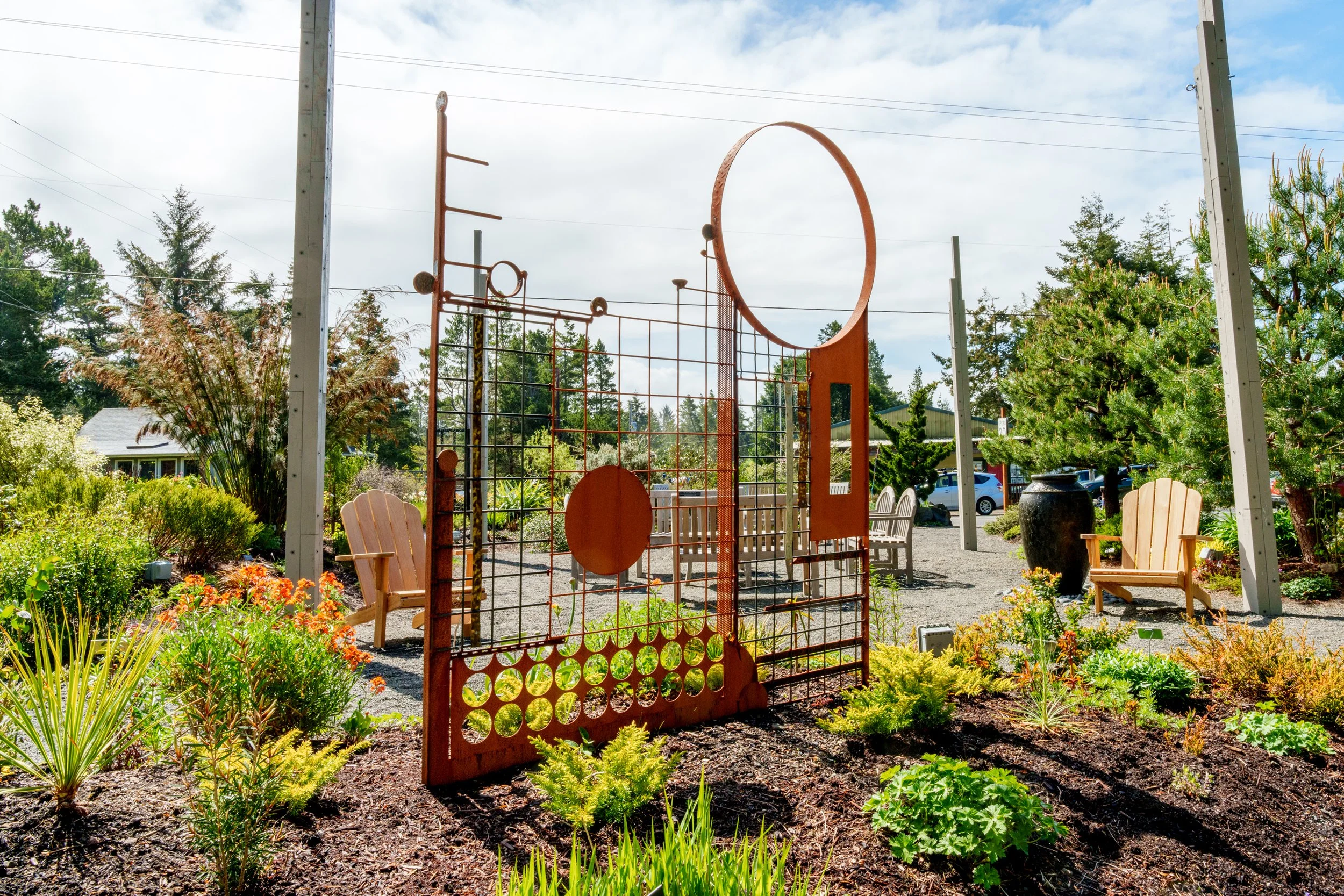 Outdoor garden with various plants, flowers, and trees, surrounded by benches and a decorative orange gate, under a partly cloudy sky.