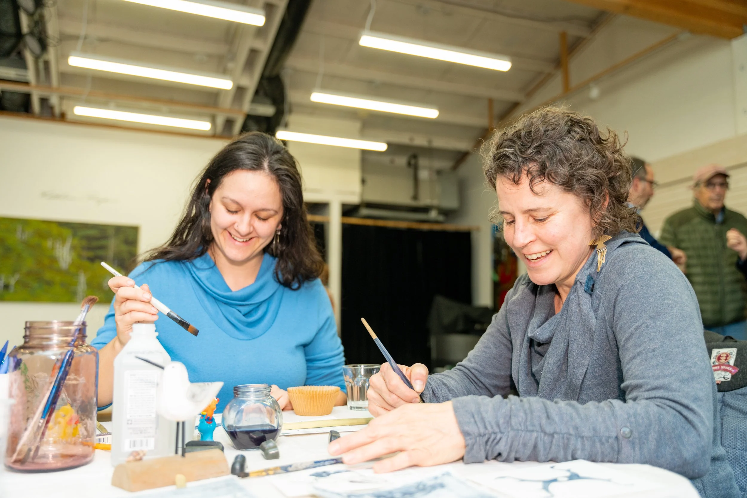 Two women are smiling and working on a project at a table, with various bottles and tools around, in an indoor setting with bright ceiling lights.