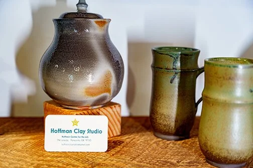 Three ceramic vases with glazed finishes on a wooden surface, one with a mottled brown and gray glaze and two with greenish glazes, with a business card for Hoffman Clay Studio in front.